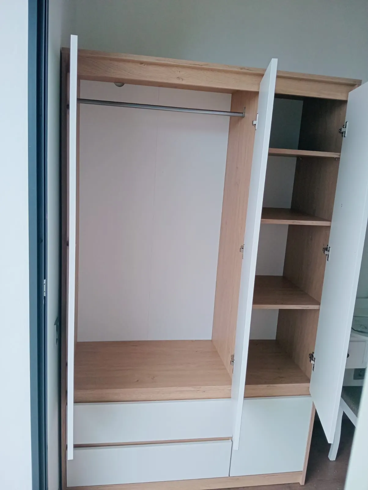 Armoire à portes blanches ouvertes sur une penderie et des étagères en bois clair, tiroirs en partie basse.