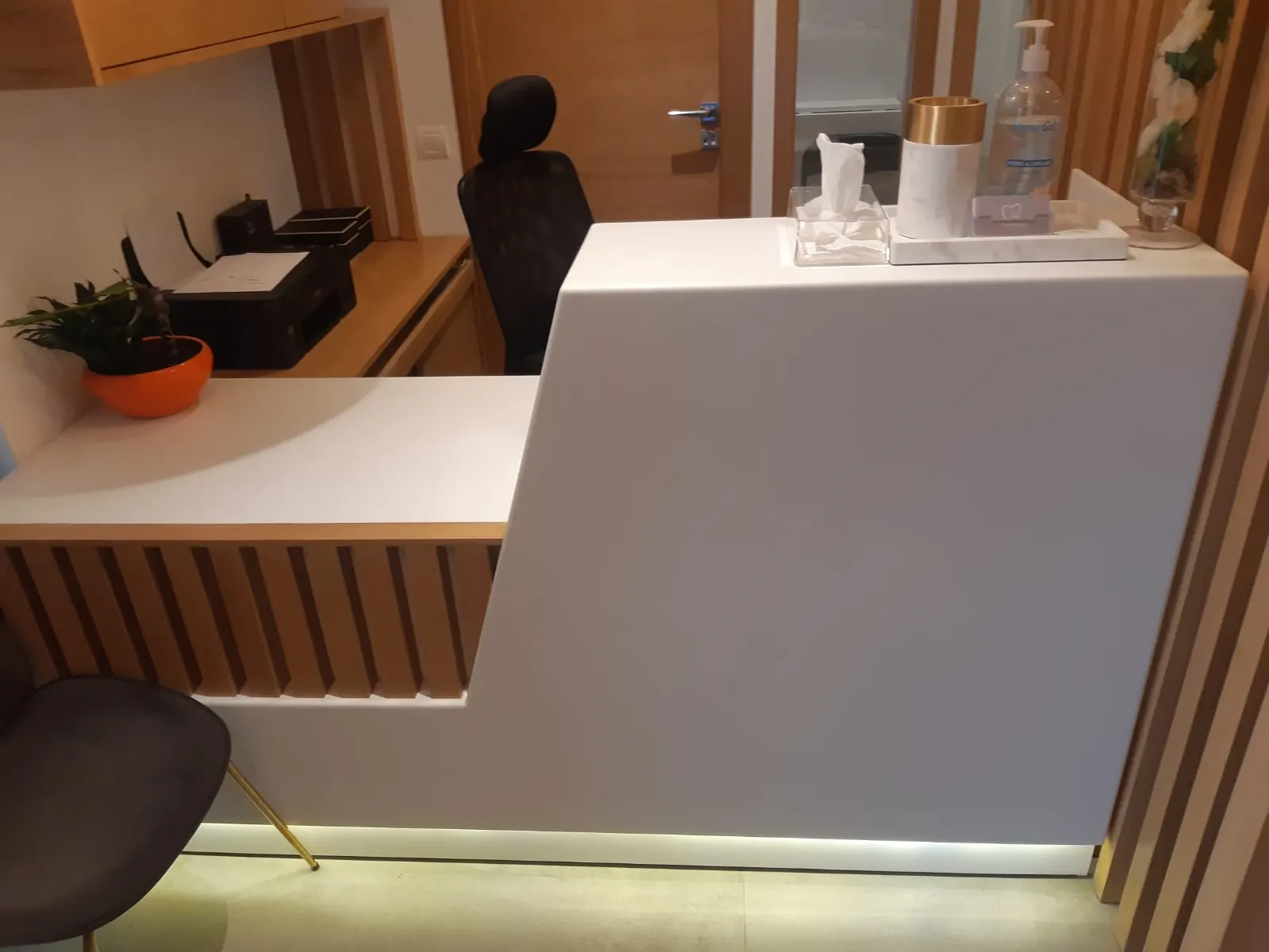 Banque d’accueil blanche à deux hauteurs, soubassement à tasseaux de bois éclairé au pied, fauteuil noir derrière.