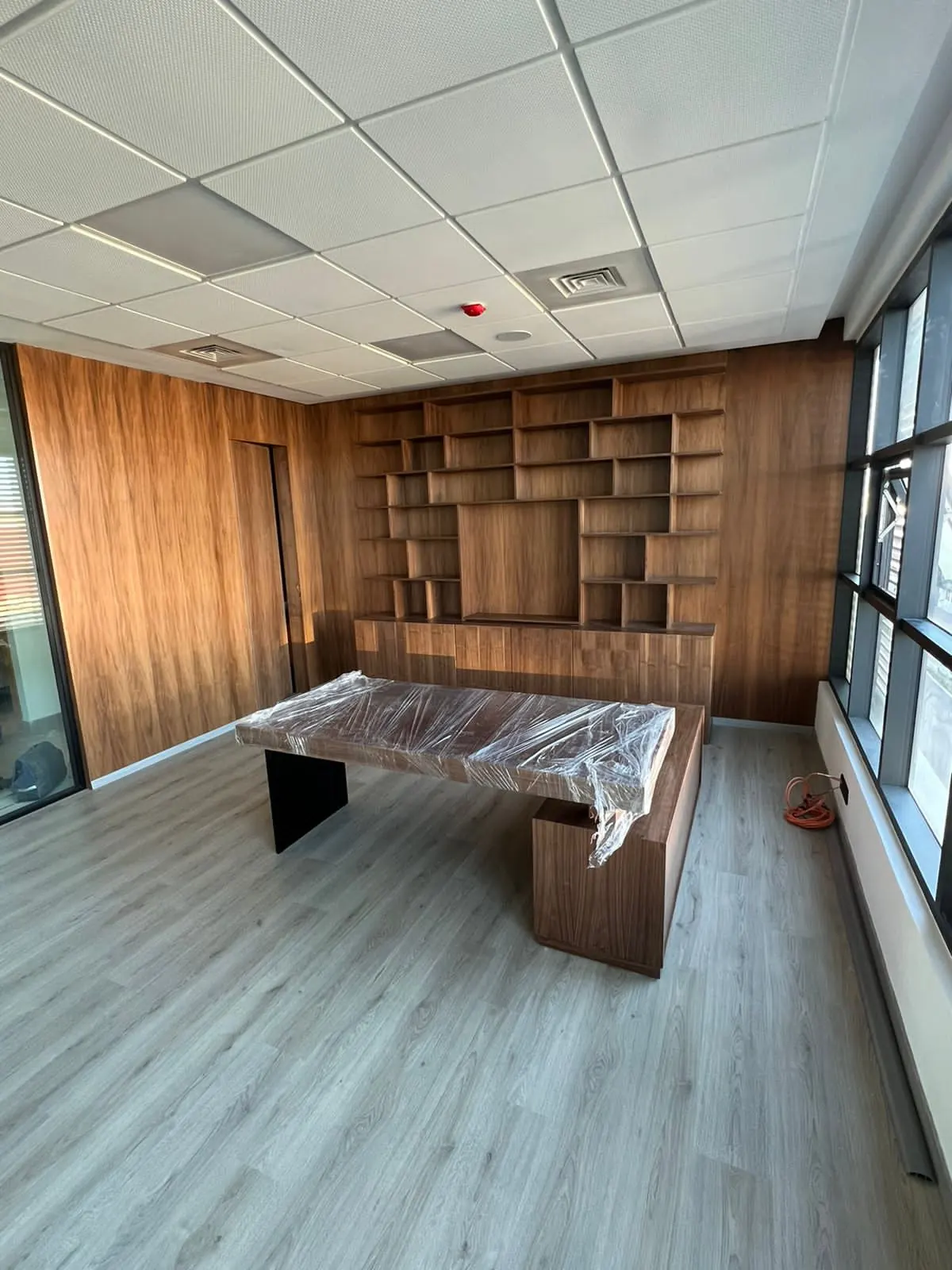 Bureau aux murs habillés de bois foncé, bibliothèque à cases au fond, bureau sous film plastique, sol à lames grises.