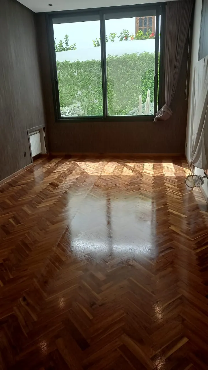 Chambre au parquet à bâtons rompus en bois foncé vitrifié, soleil d’une baie vitrée en reflet sur le sol.