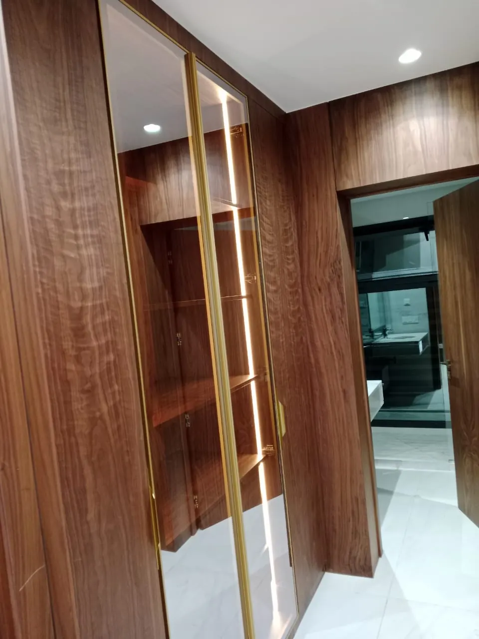 Dressing habillé de bois foncé veiné, porte vitrée à profils dorés et éclairage vertical, salle de bain ouverte au fond.