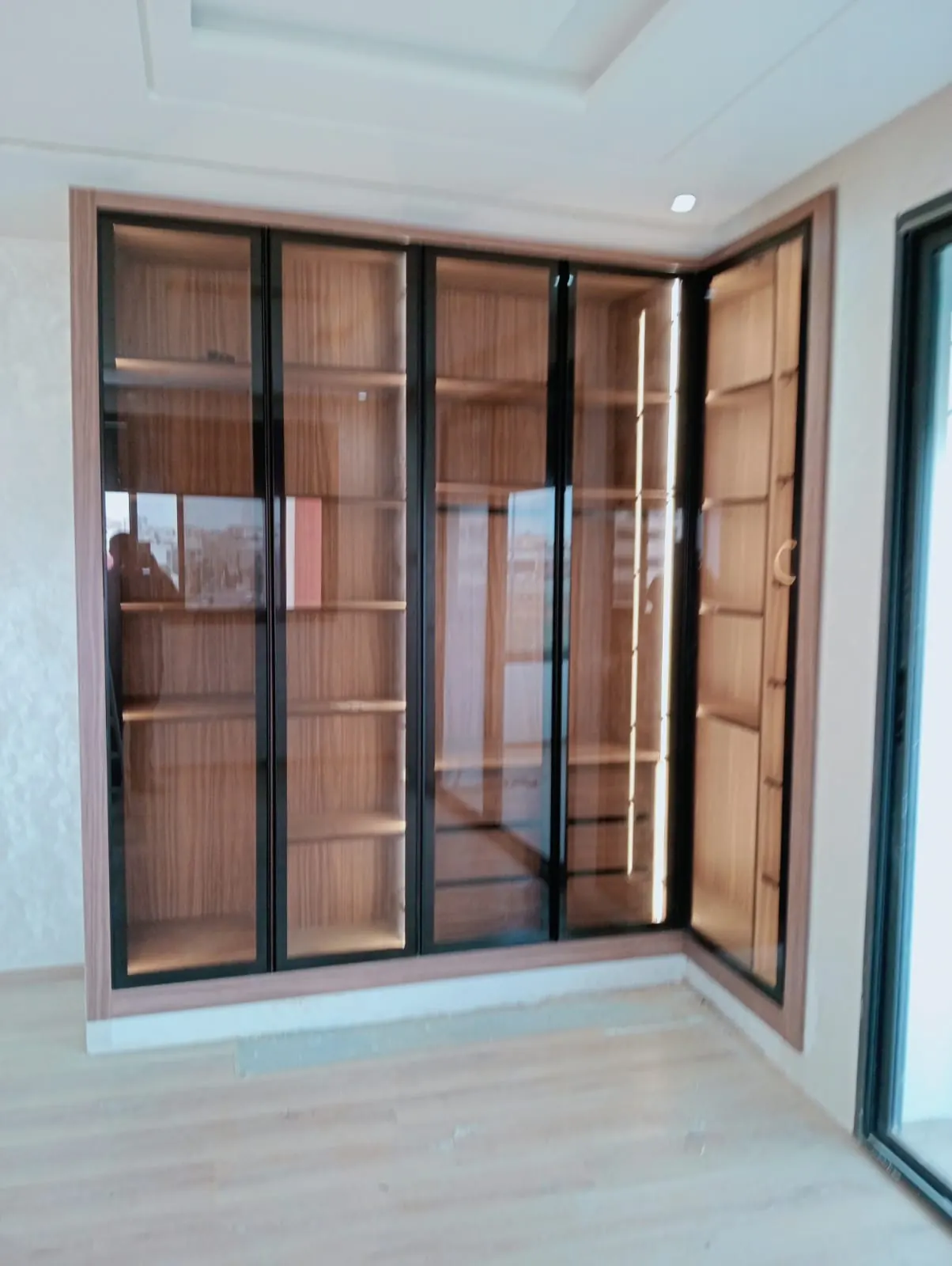 Dressing d’angle sur mesure à portes vitrées cadrées de noir, étagères en bois éclairées par des bandeaux lumineux.