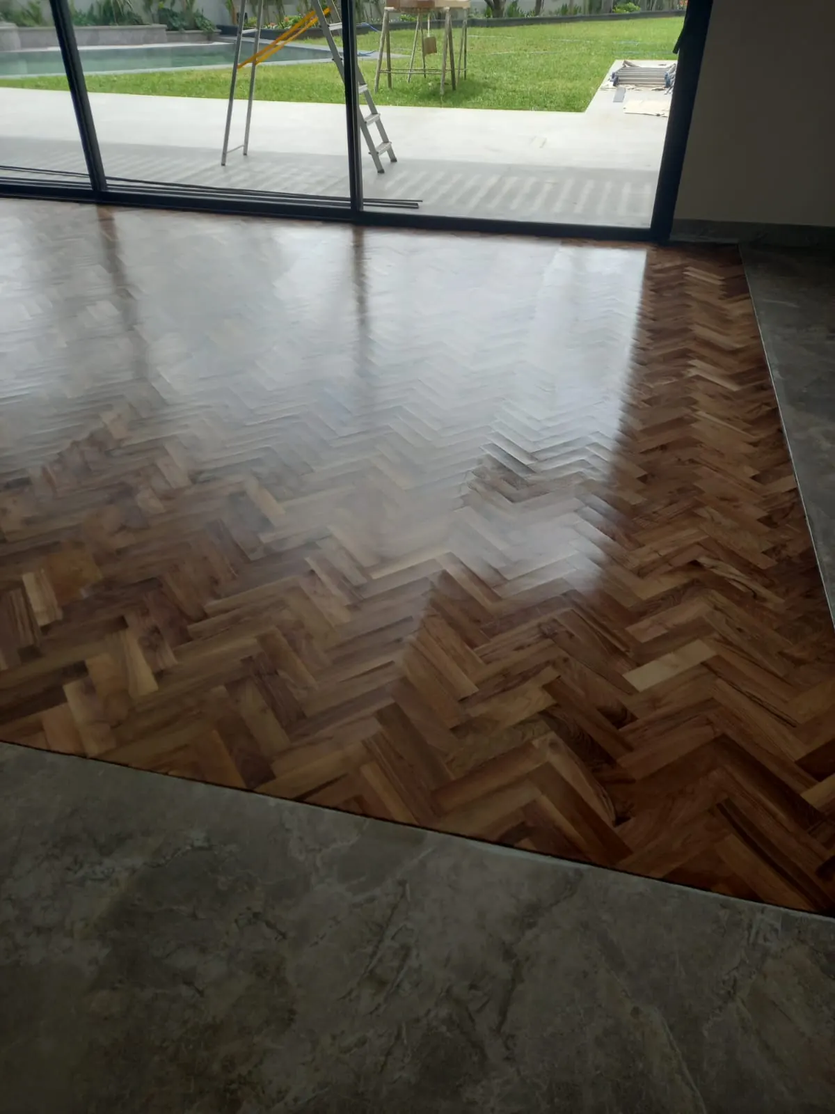 Parquet à bâtons rompus en bois foncé vitrifié, bordé d’un sol en marbre gris, devant une baie ouverte sur un jardin.