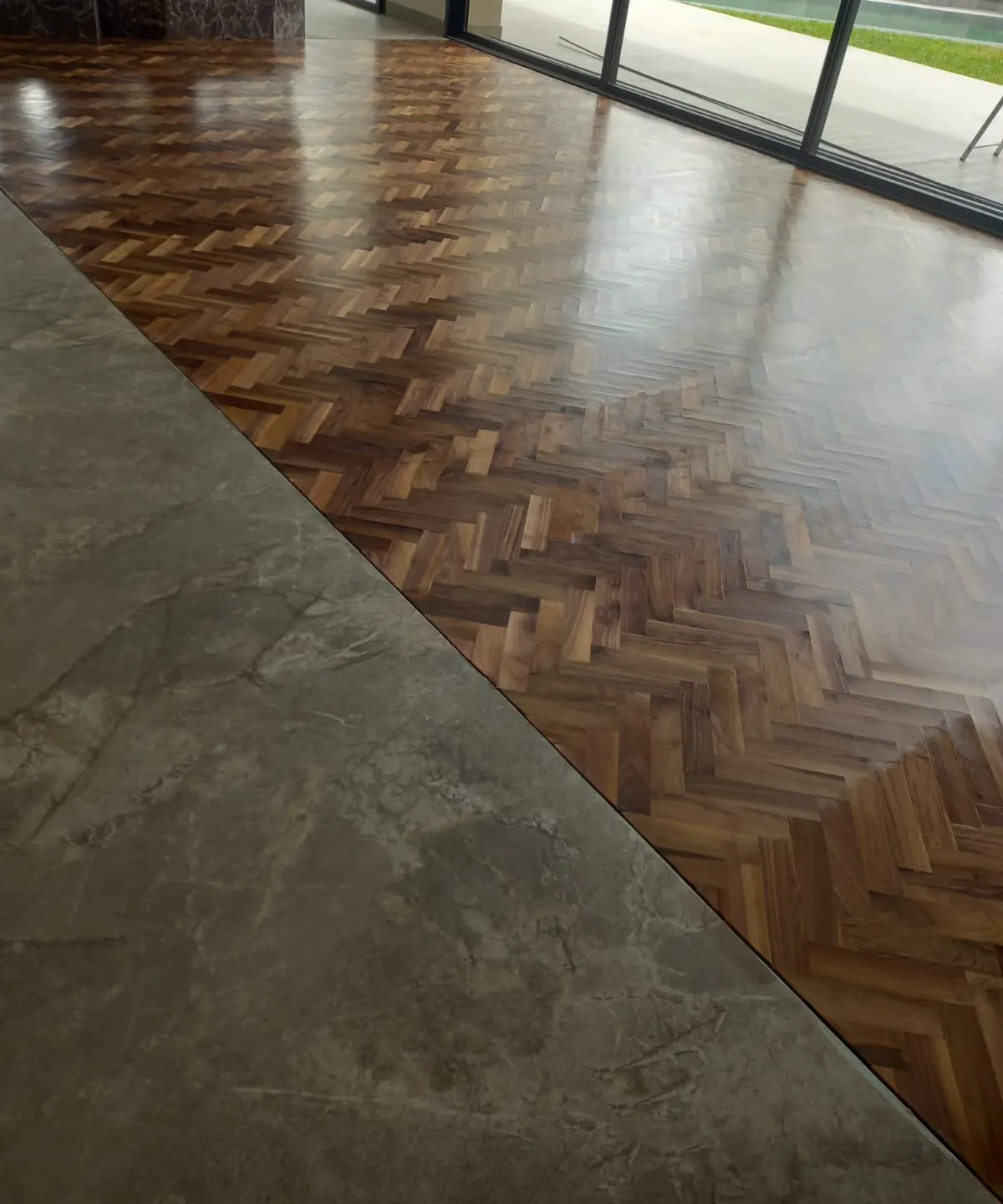 Parquet à bâtons rompus en bois foncé vitrifié raccordé en biais à un sol en marbre gris, baies vitrées sur une piscine.