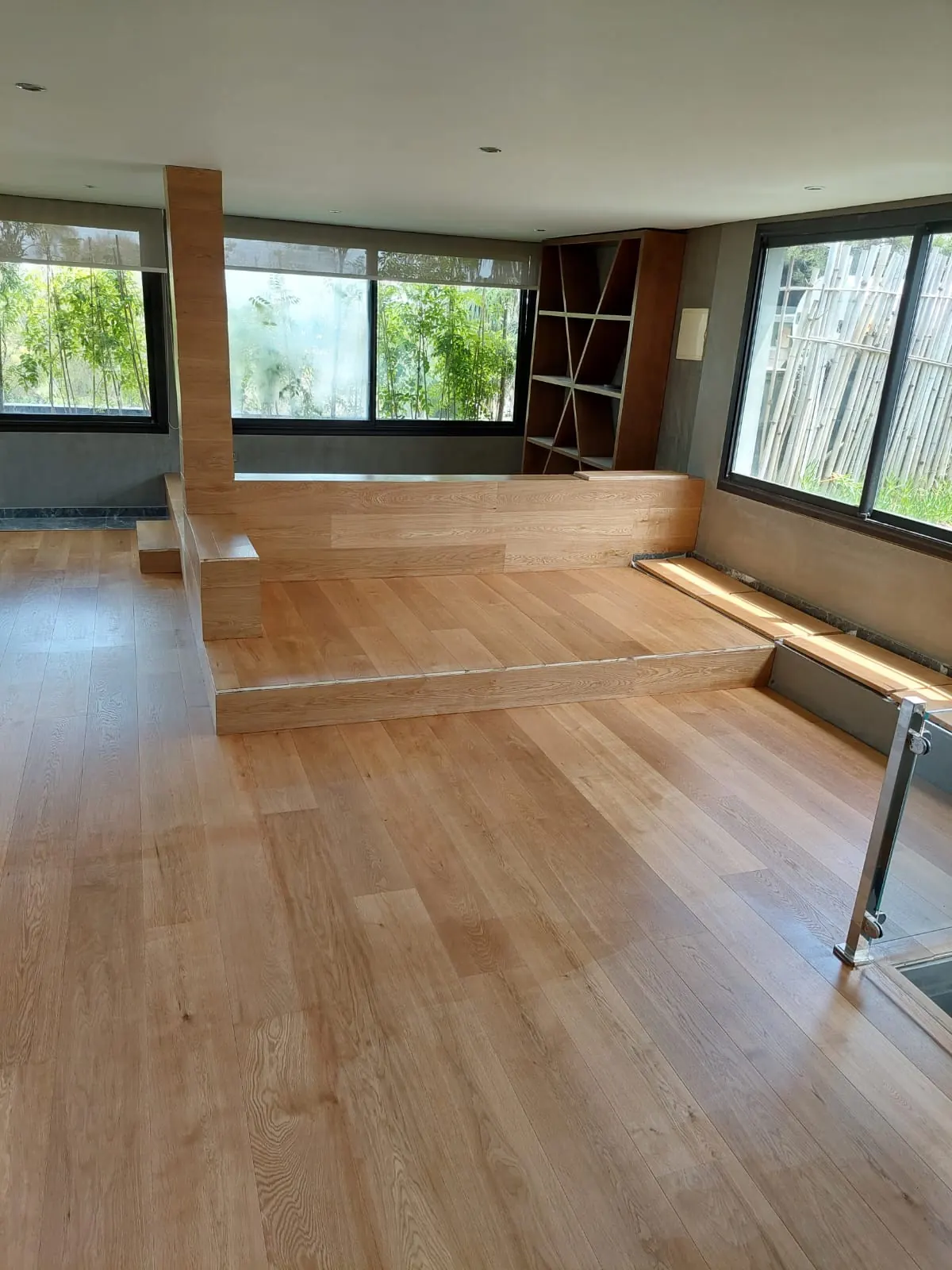Séjour au parquet à lames larges en bois clair, estrade et banquette habillées du même bois, étagères et grandes fenêtres.