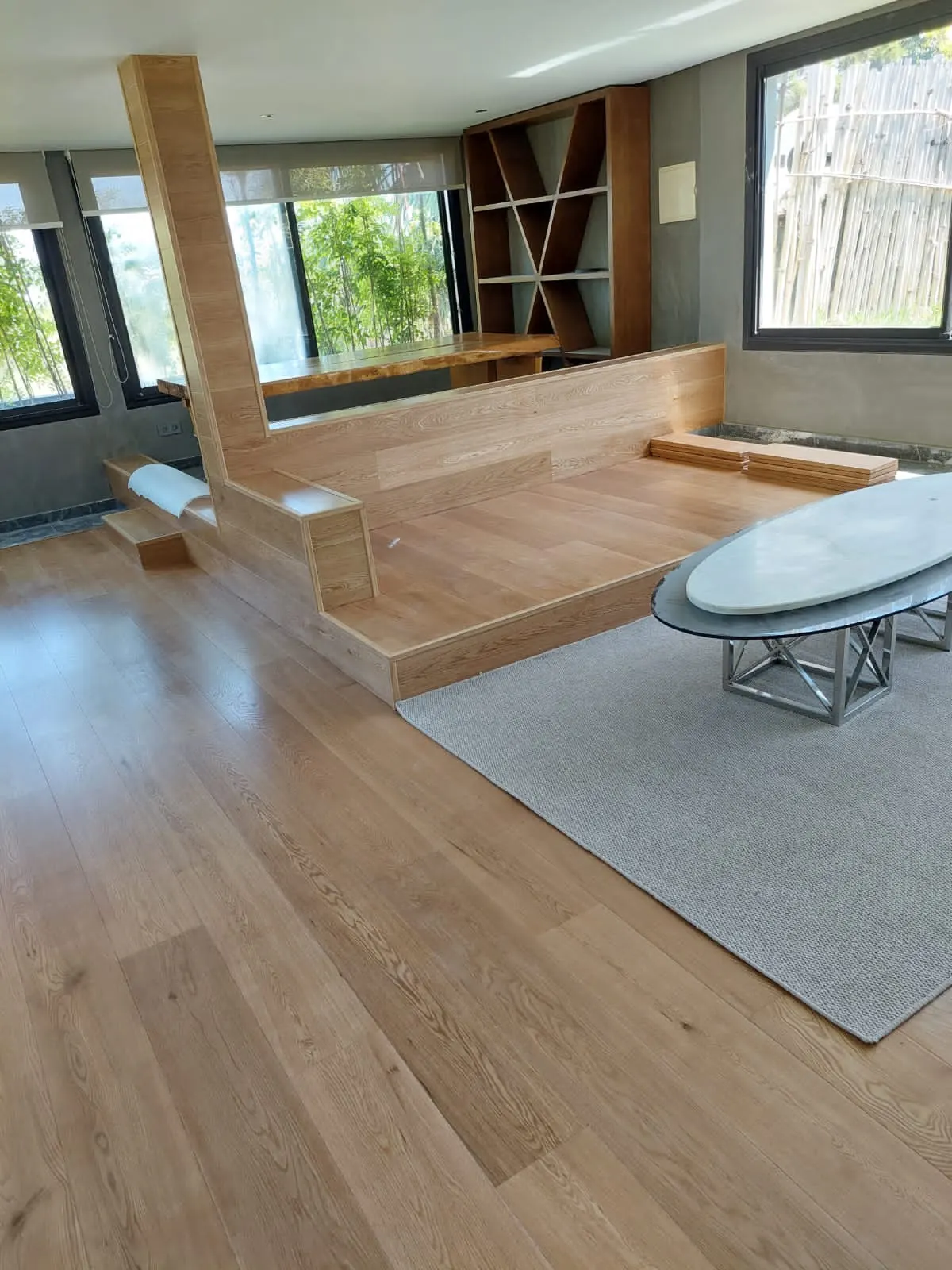Séjour au parquet à lames larges en bois clair, estrade en bois, tapis gris et table basse ovale en marbre.