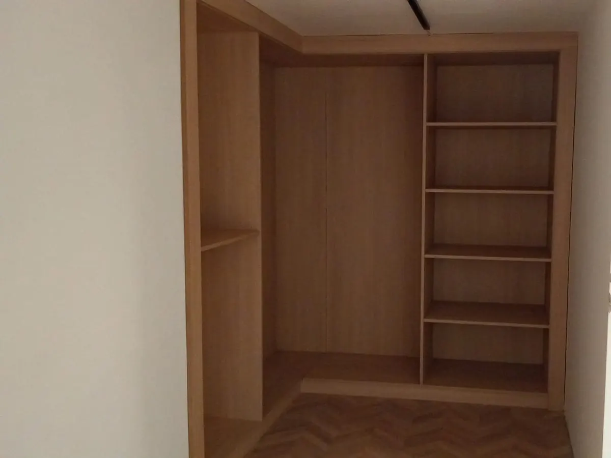 Placard d’angle en bois clair encastré dans une niche, encore sans portes : grand volume à gauche, colonne d’étagères à droite, parquet au sol.