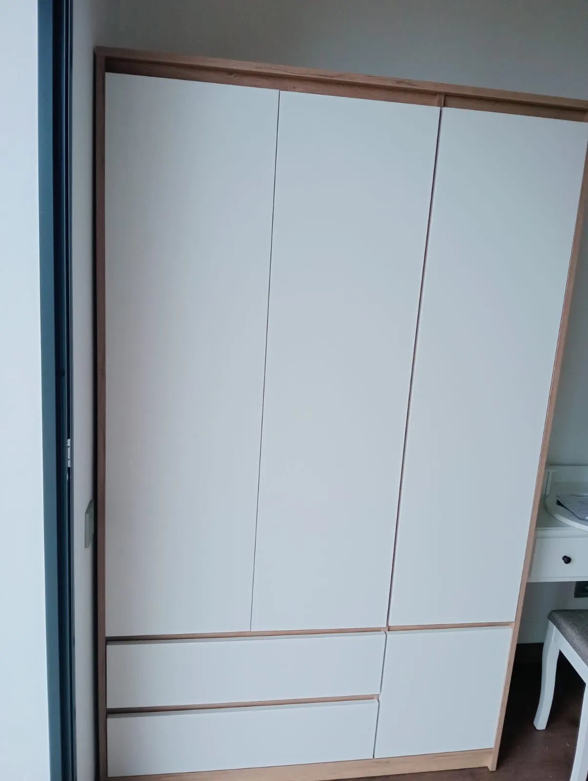Placard de chambre fermé : trois portes et trois tiroirs à façades blanches lisses dans un cadre en bois clair, une coiffeuse blanche à côté.