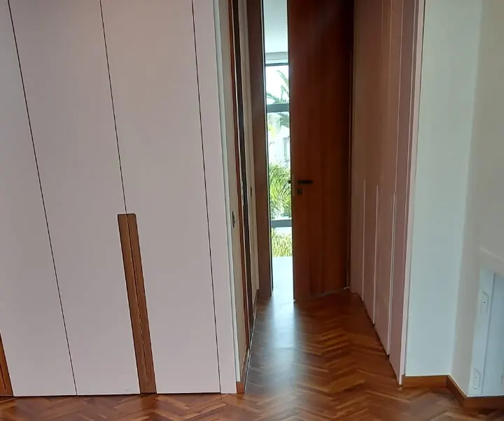 Placards de couloir à façades blanches et longues poignées en bois encastrées, porte ouverte au fond, parquet en bois foncé au sol.