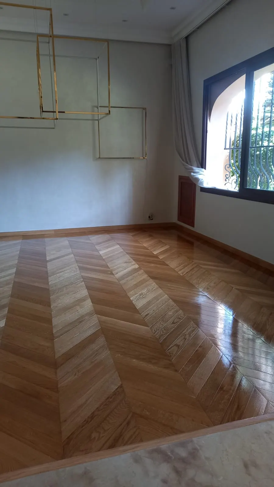 Salon au parquet en point de Hongrie en bois clair vitrifié, suspension en cadres dorés et fenêtre à grille en fer forgé.