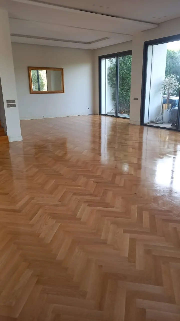 Grand séjour vide au parquet à bâtons rompus en bois clair vitrifié, baies vitrées sur un jardin et miroir au mur.