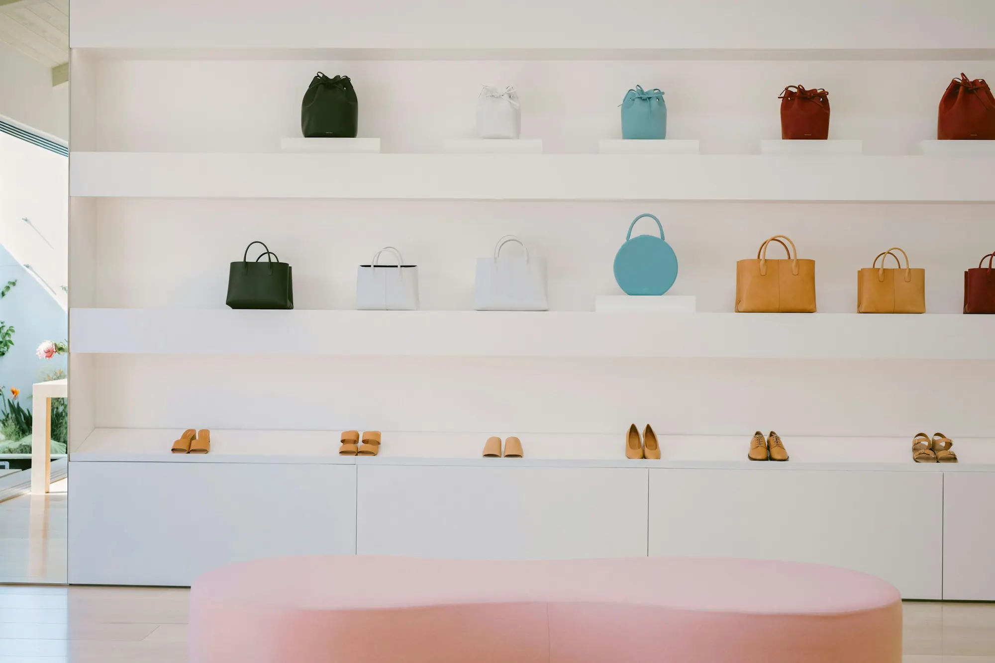 Agencement de boutique blanche : sacs et chaussures alignés sur des étagères murales, banquette rose au premier plan.