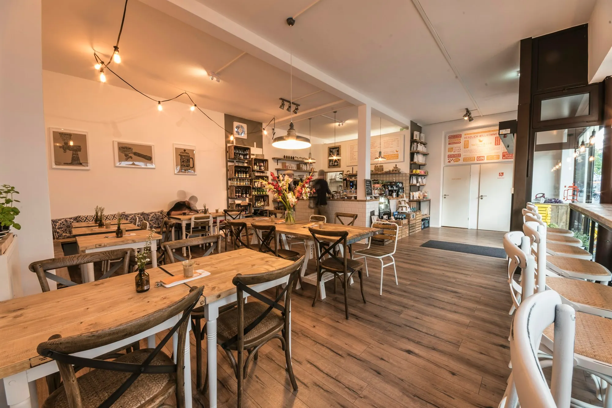 Agencement de café chaleureux : tables en bois brut, chaises bistrot, sol en bois foncé, guirlande d’ampoules.