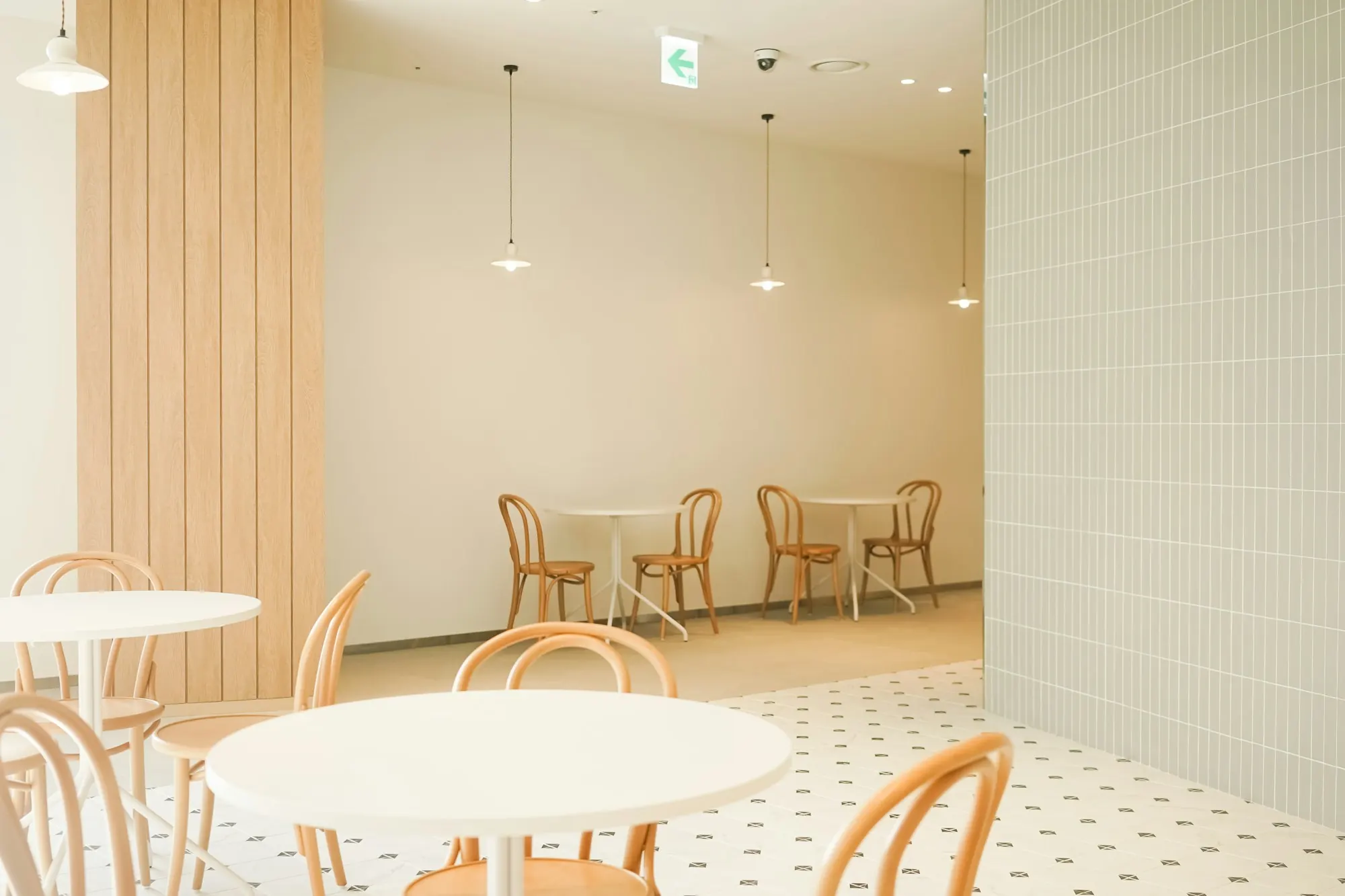 Agencement de café clair : tables rondes blanches, chaises bistrot en bois courbé, carrelage à motifs.