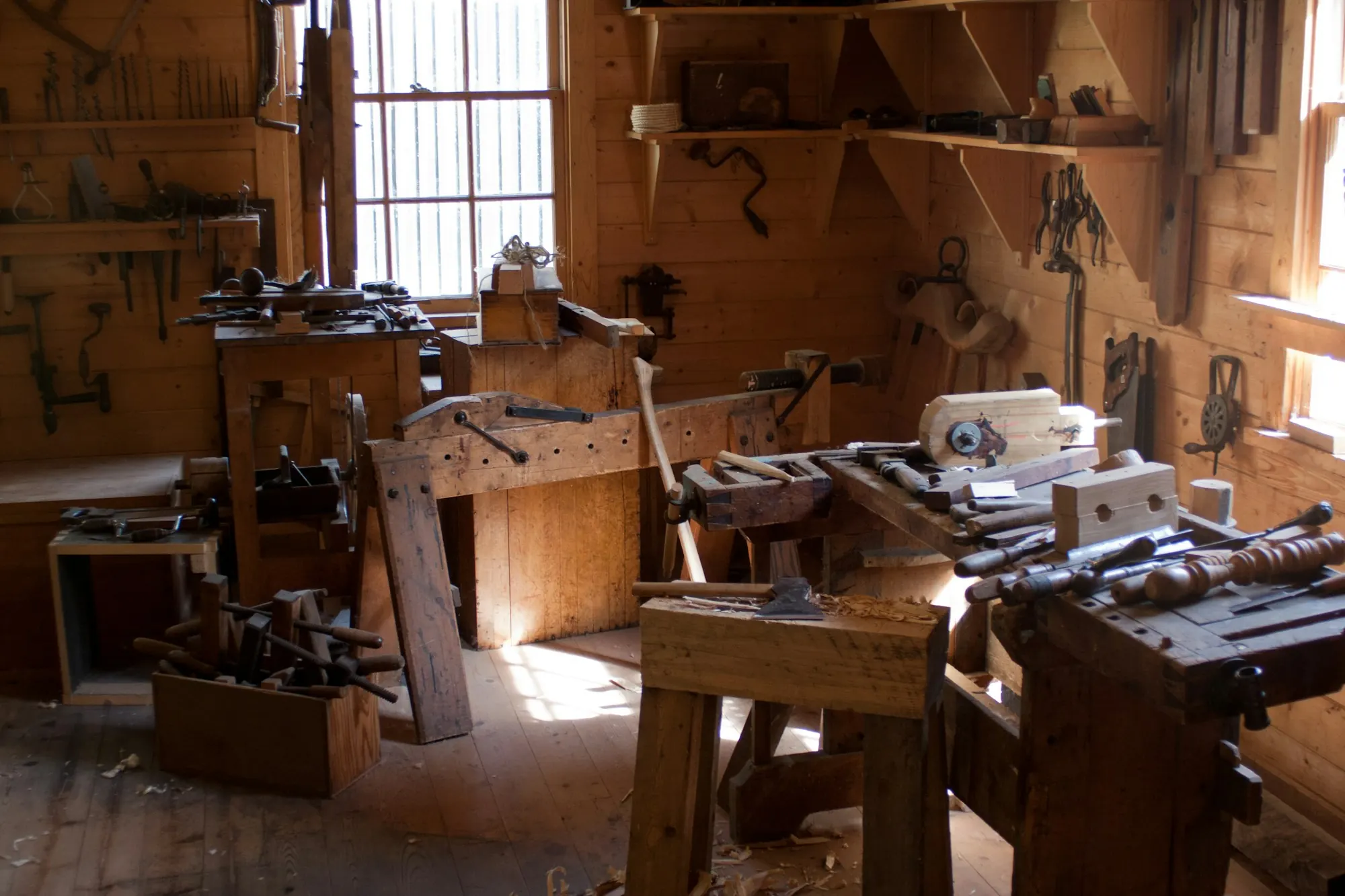 Atelier de menuiserie ancien : établis en bois, outils à main aux murs, lumière d’une fenêtre.