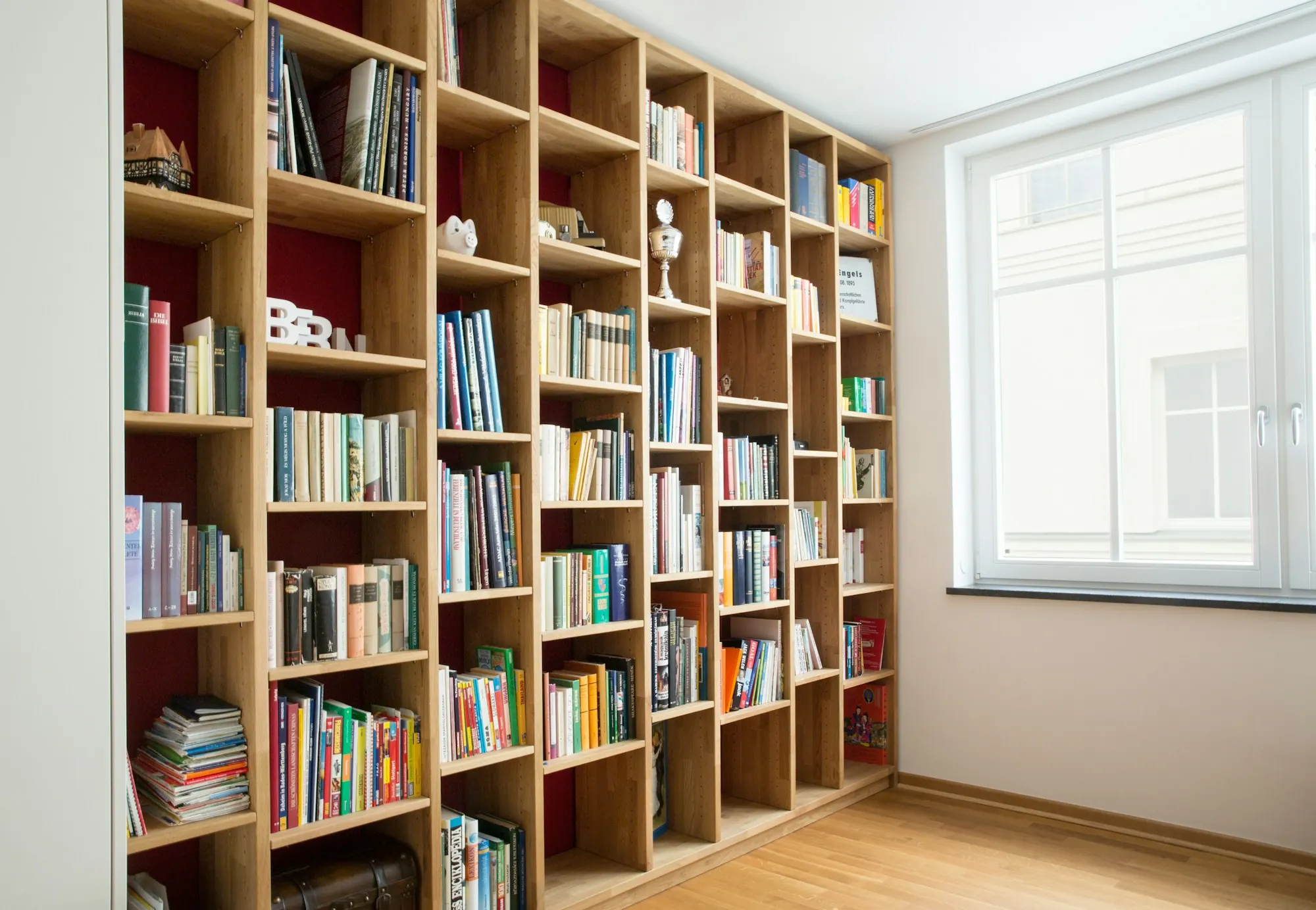 Bibliothèque en bois clair du sol au plafond sur fond rouge sombre, garnie de livres, près d’une fenêtre.