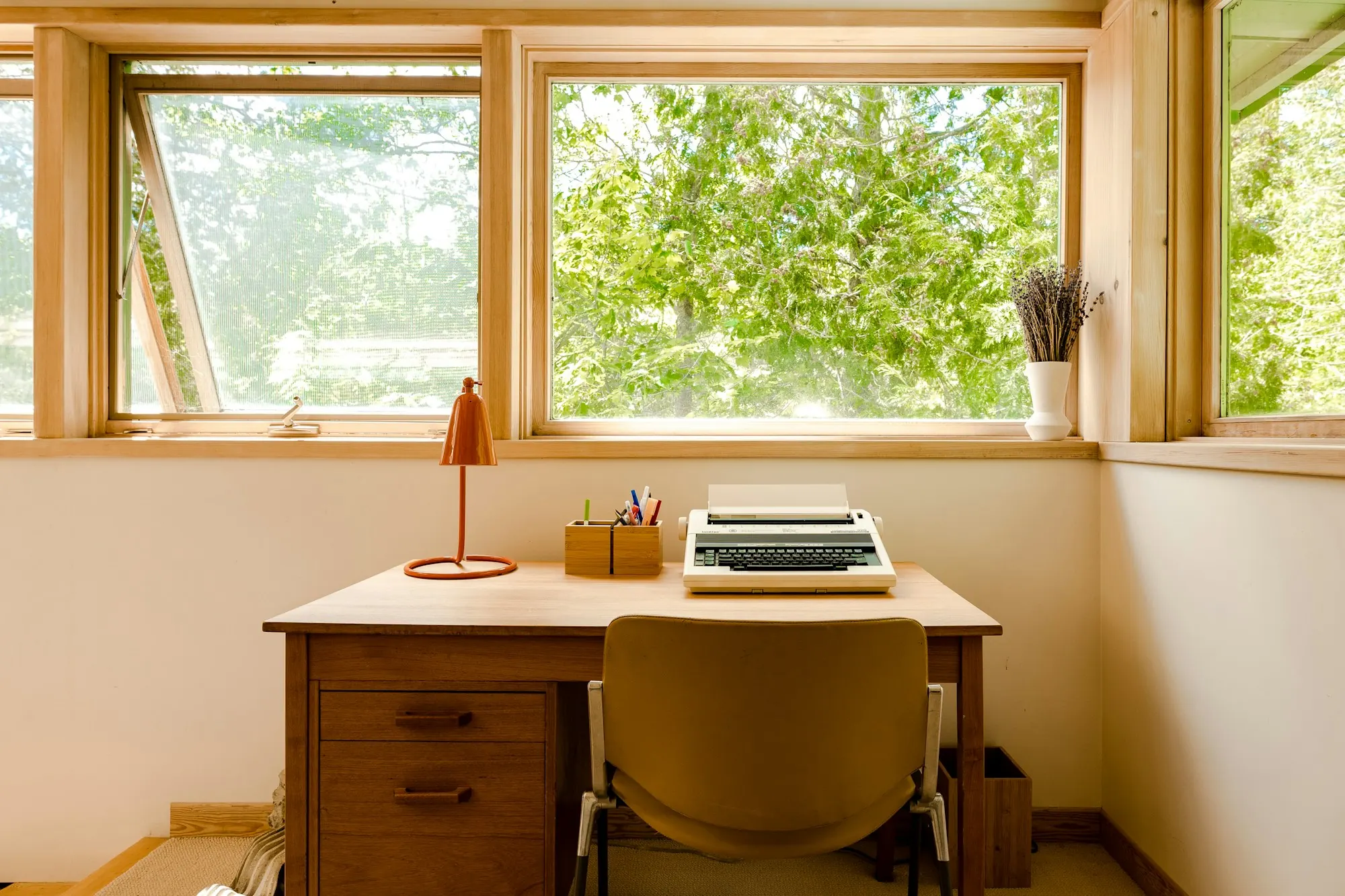 Bureau en bois devant une large fenêtre à cadres de bois ouverte sur des arbres, machine à écrire et lampe orange.