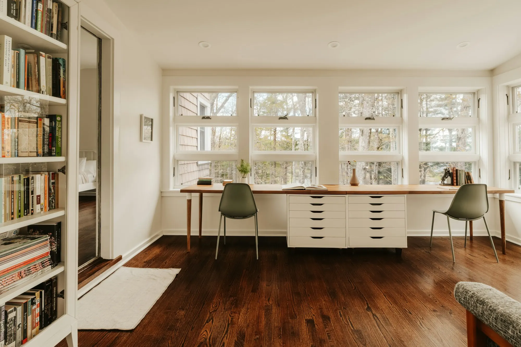 Bureau sur parquet foncé à lames étroites : long plan de travail sous une rangée de fenêtres, bibliothèque pleine hauteur.