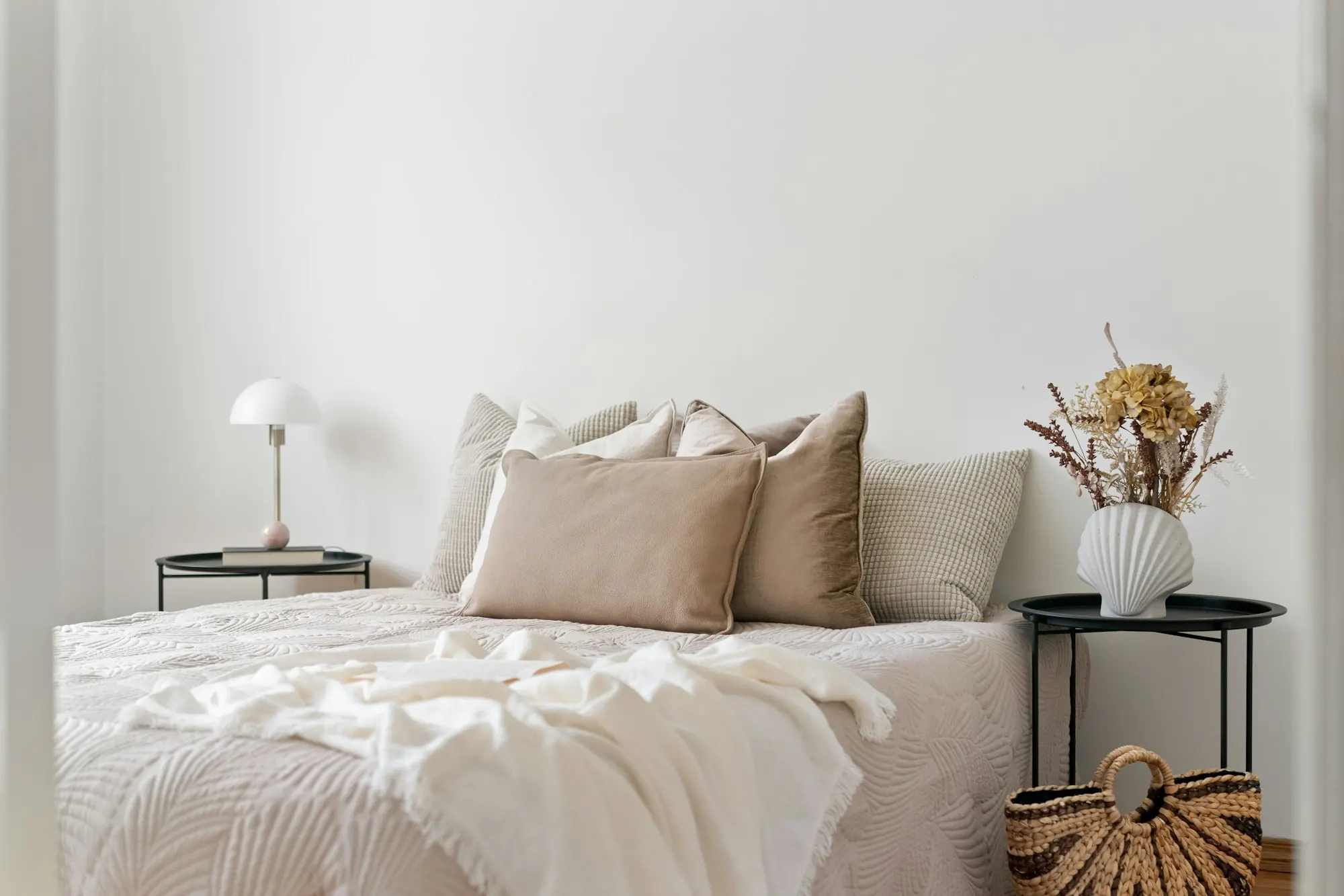 Chambre neutre : lit à coussins beiges et couvre-lit blanc, tables de chevet noires, mur blanc.