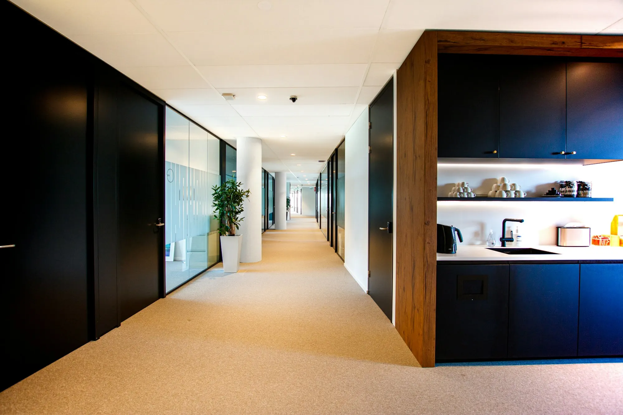 Couloir de bureaux sur moquette beige : portes noires, parois vitrées, kitchenette encadrée de bois.