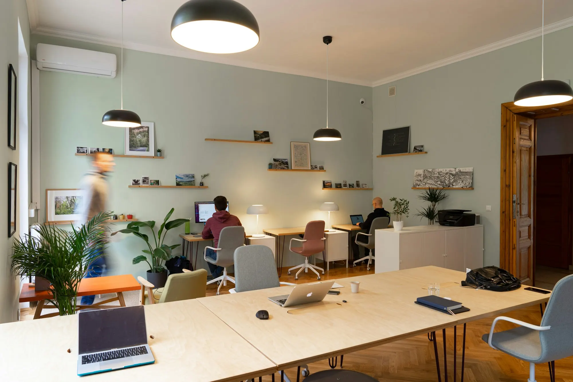 Espace de coworking sur parquet à bâtons rompus : grandes tables en contreplaqué, suspensions noires, murs vert d’eau.