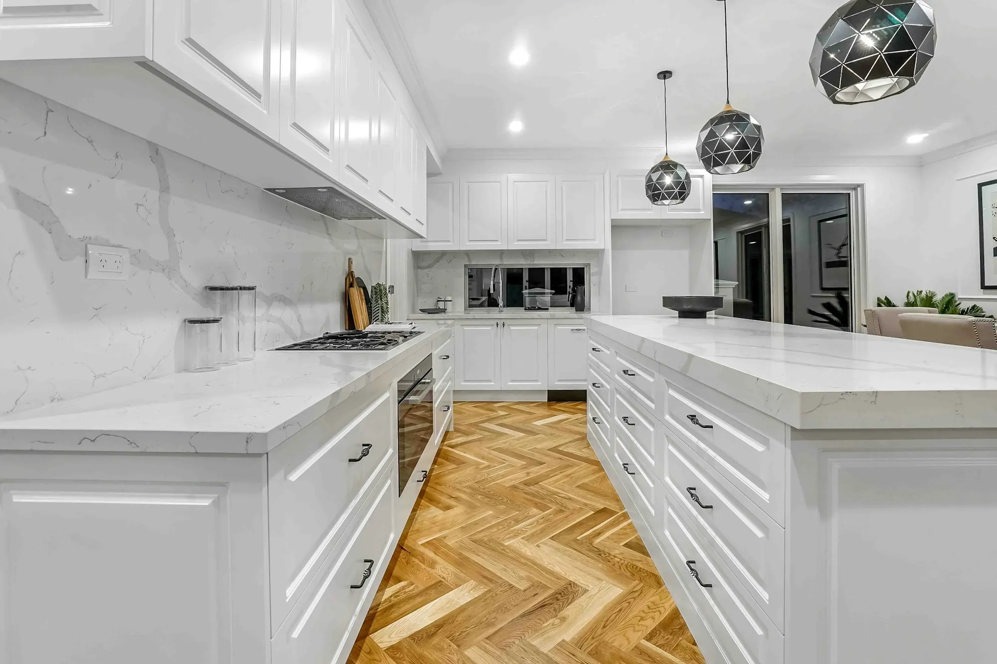 Cuisine blanche à moulures : îlot et plans en pierre veinée, parquet blond à bâtons rompus, suspensions à facettes.