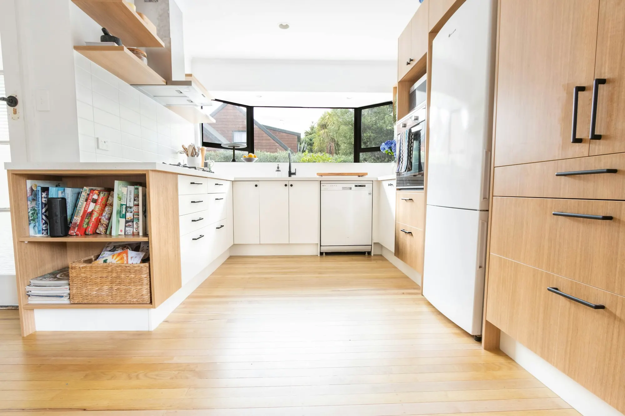 Cuisine en U : façades blanches et colonnes en bois clair, niche à livres, parquet blond, fenêtre d’angle.