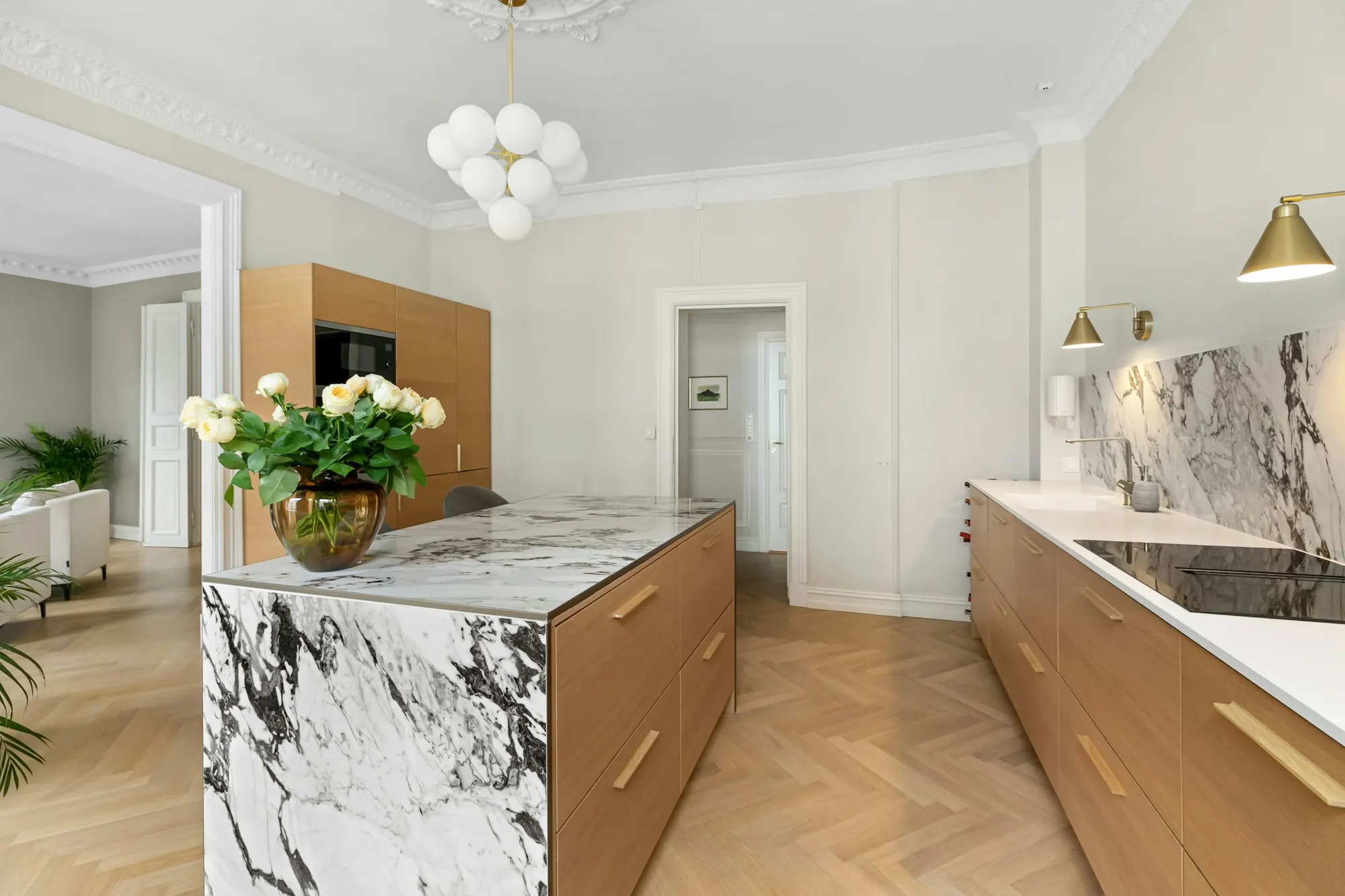Cuisine sur parquet clair à bâtons rompus : îlot en marbre veiné, façades en bois, moulures et suspension à globes.