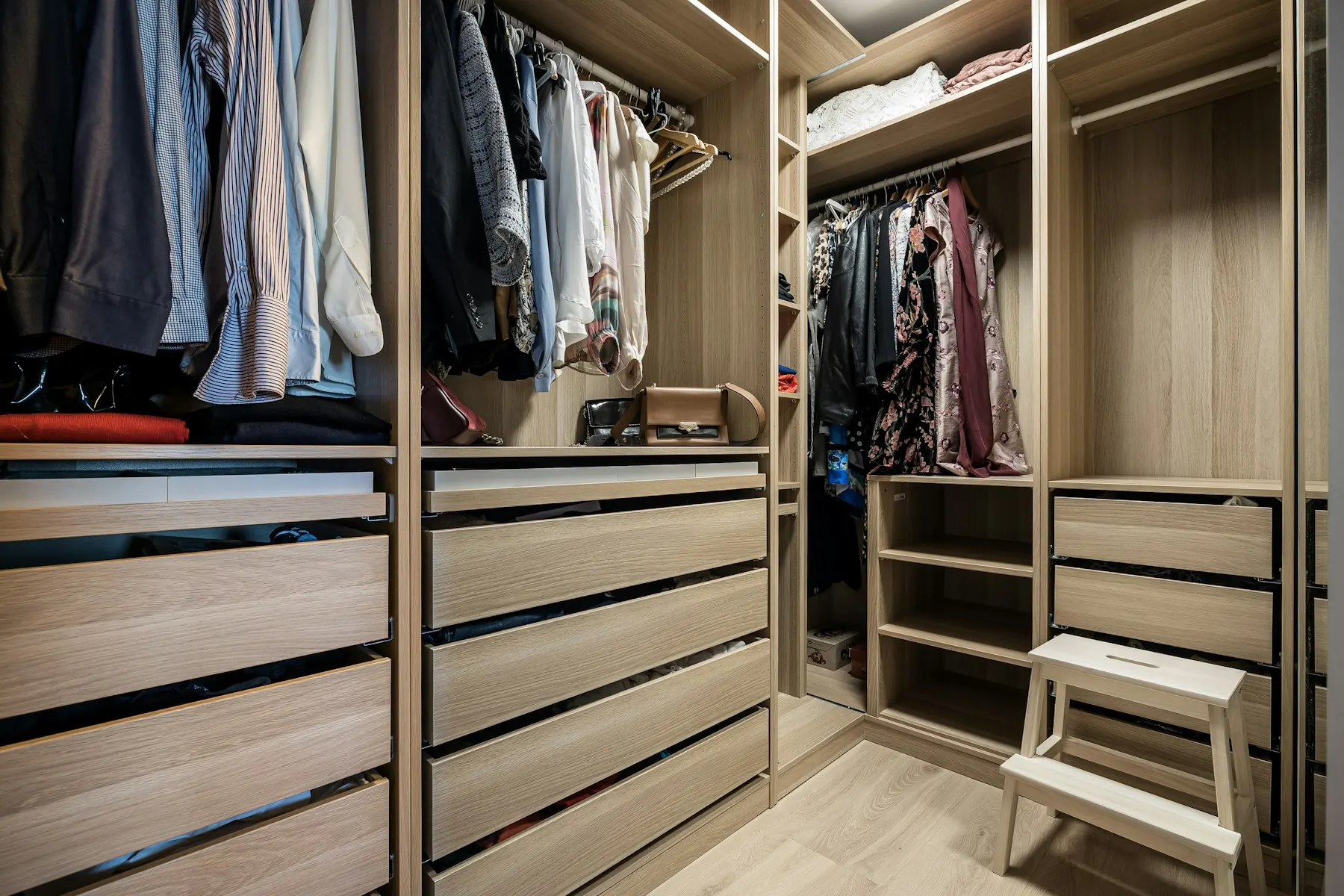 Dressing sur mesure en bois clair : penderies garnies, tiroirs, étagères d’angle et petit marchepied blanc.