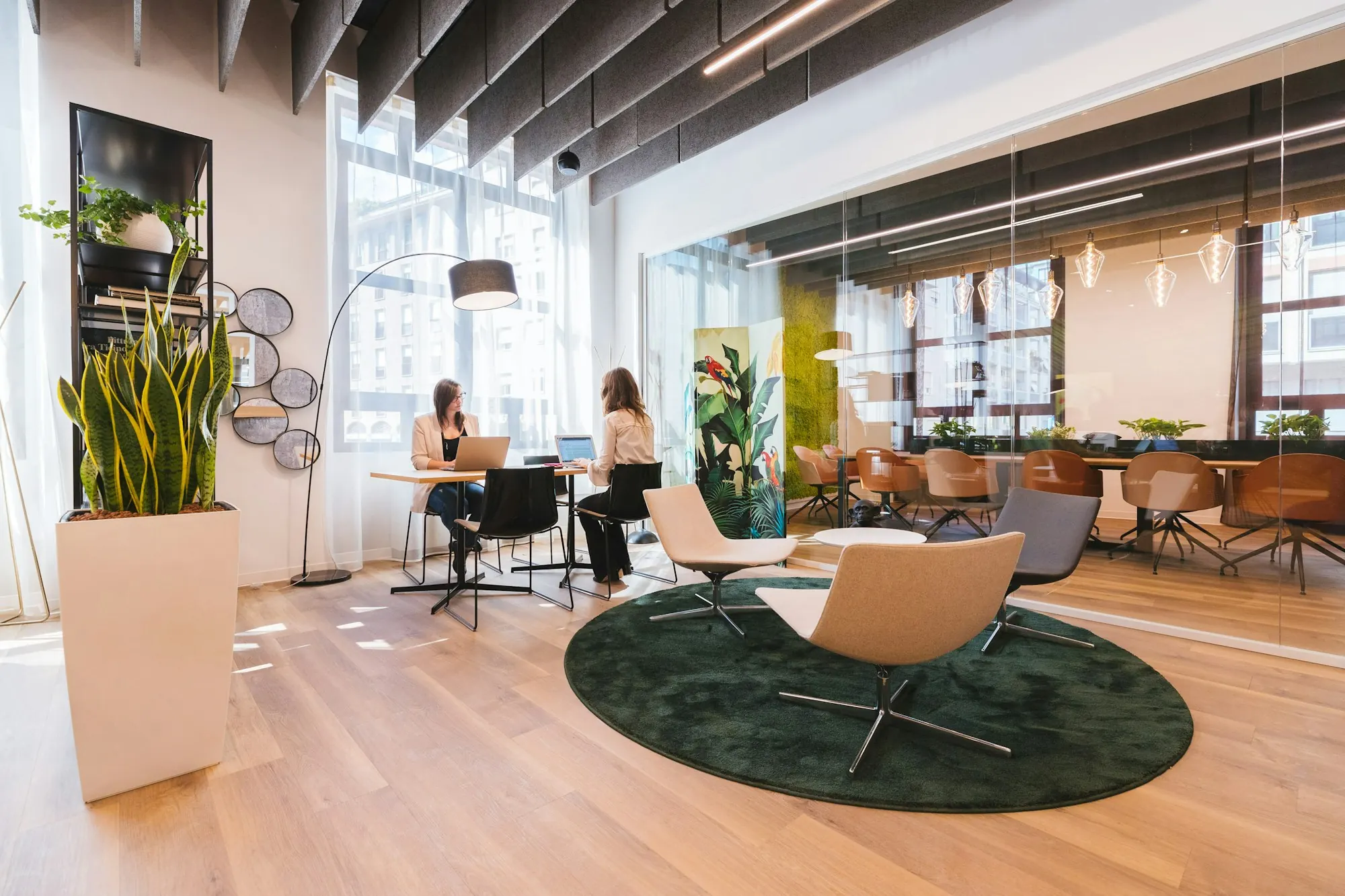 Plateau de bureaux sur parquet clair : fauteuils autour d’un tapis rond vert, panneaux suspendus au plafond, salle vitrée.