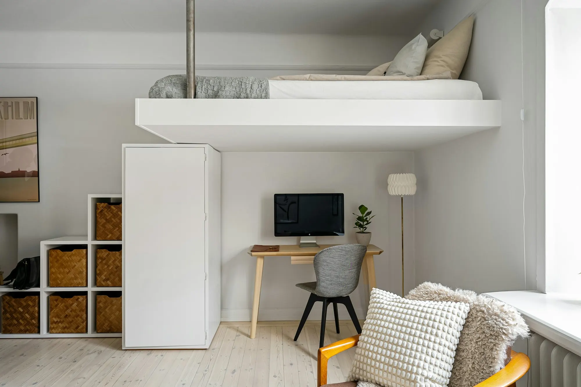 Lit en mezzanine blanc au-dessus d’un bureau en bois clair, penderie blanche et casiers à paniers tressés.