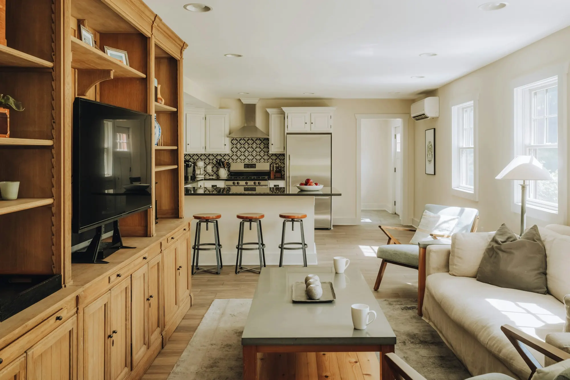 Séjour ouvert sur la cuisine : meuble TV et bibliothèque en bois intégrés au mur, canapé clair, sol en bois.