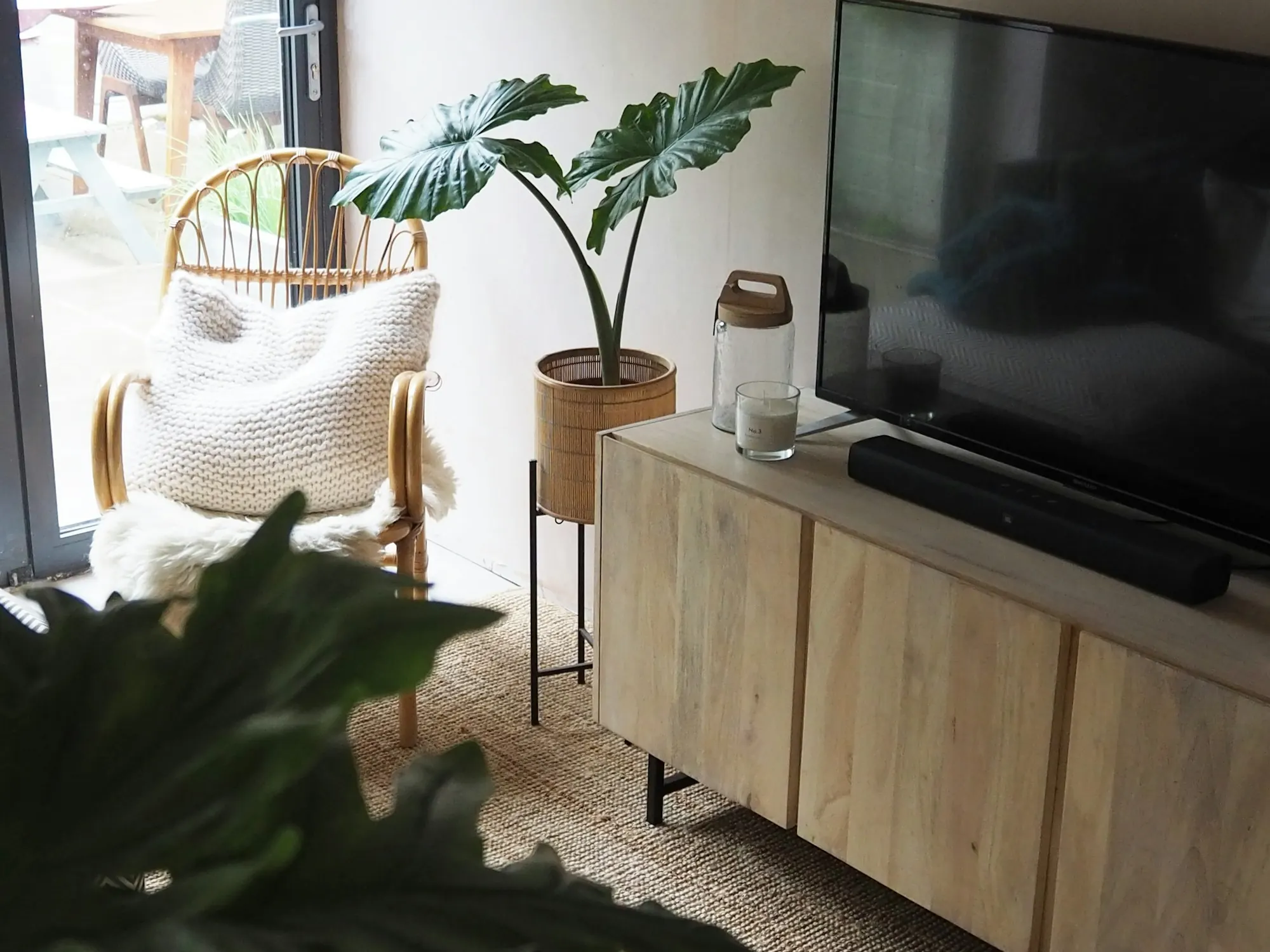 Meuble TV en bois clair à façades lisses, fauteuil en rotin et plante en pot sur un tapis de jute.