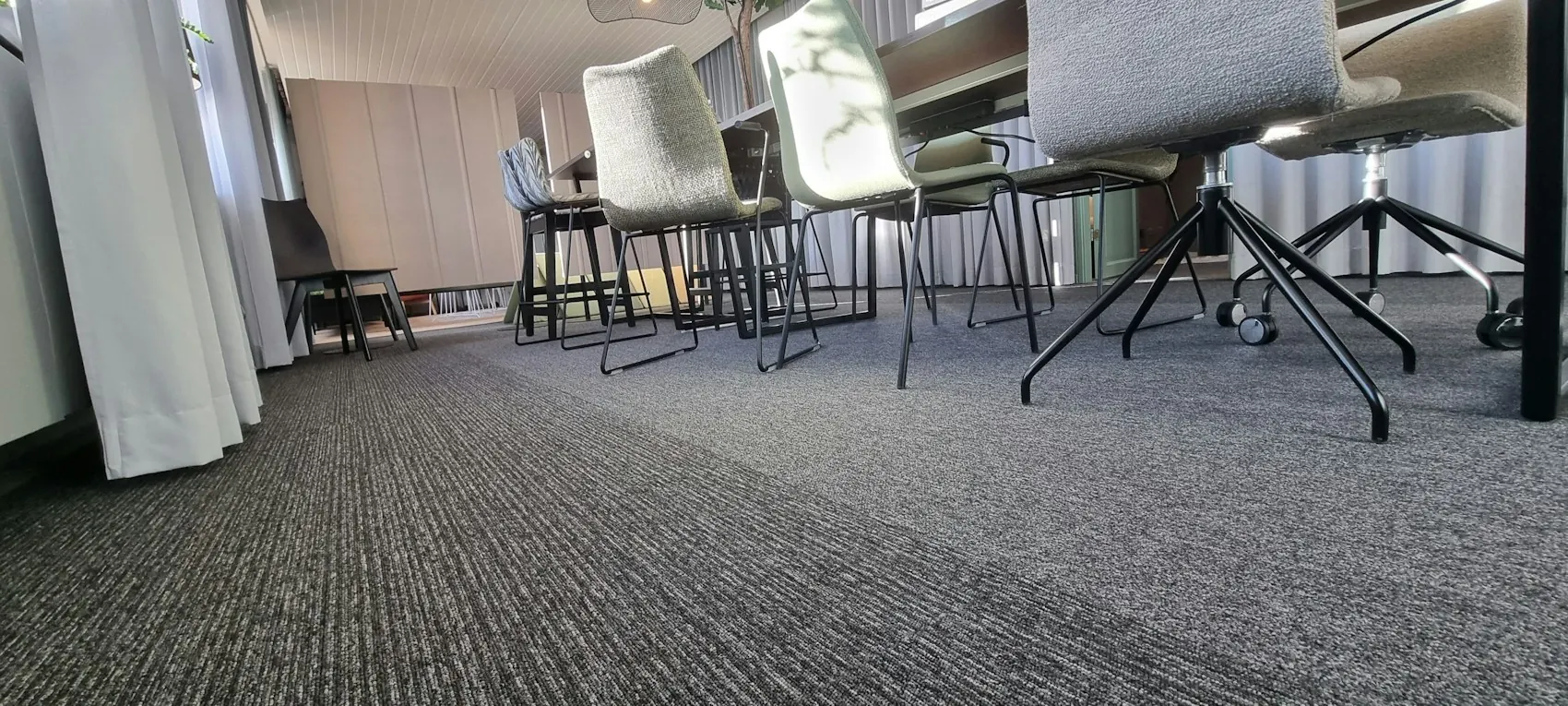 Moquette grise de bureau vue au ras du sol : piètements de chaises et de tables, voilages blancs.