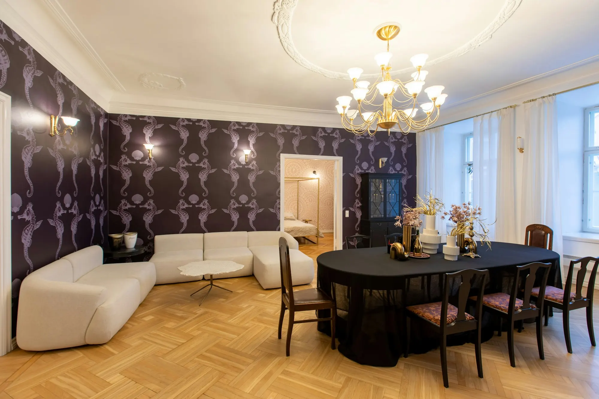 Salle à manger sous moulures : papier peint à grand motif mauve sur fond sombre, lustre doré, table ronde nappée de noir, parquet à bâtons rompus.