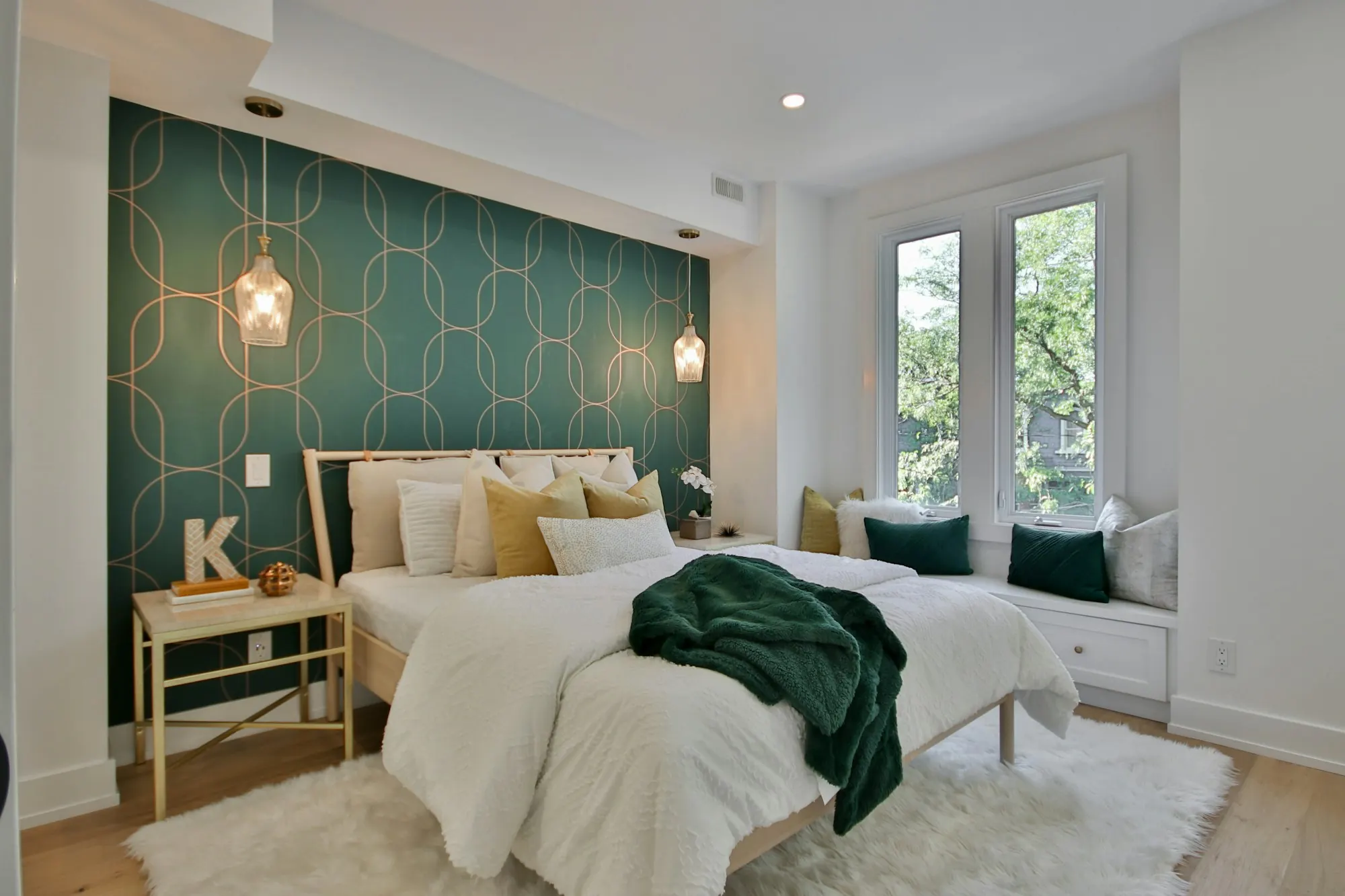 Chambre : papier peint vert sombre à arabesques dorées derrière le lit, suspensions en verre, banquette sous la fenêtre.