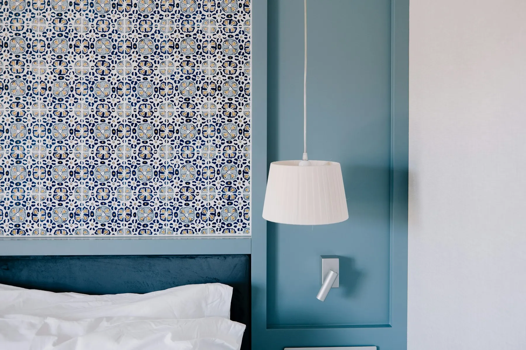 Papier peint à motif de carreaux bleus et blancs au-dessus d’une tête de lit, encadrement bleu canard, abat-jour plissé.