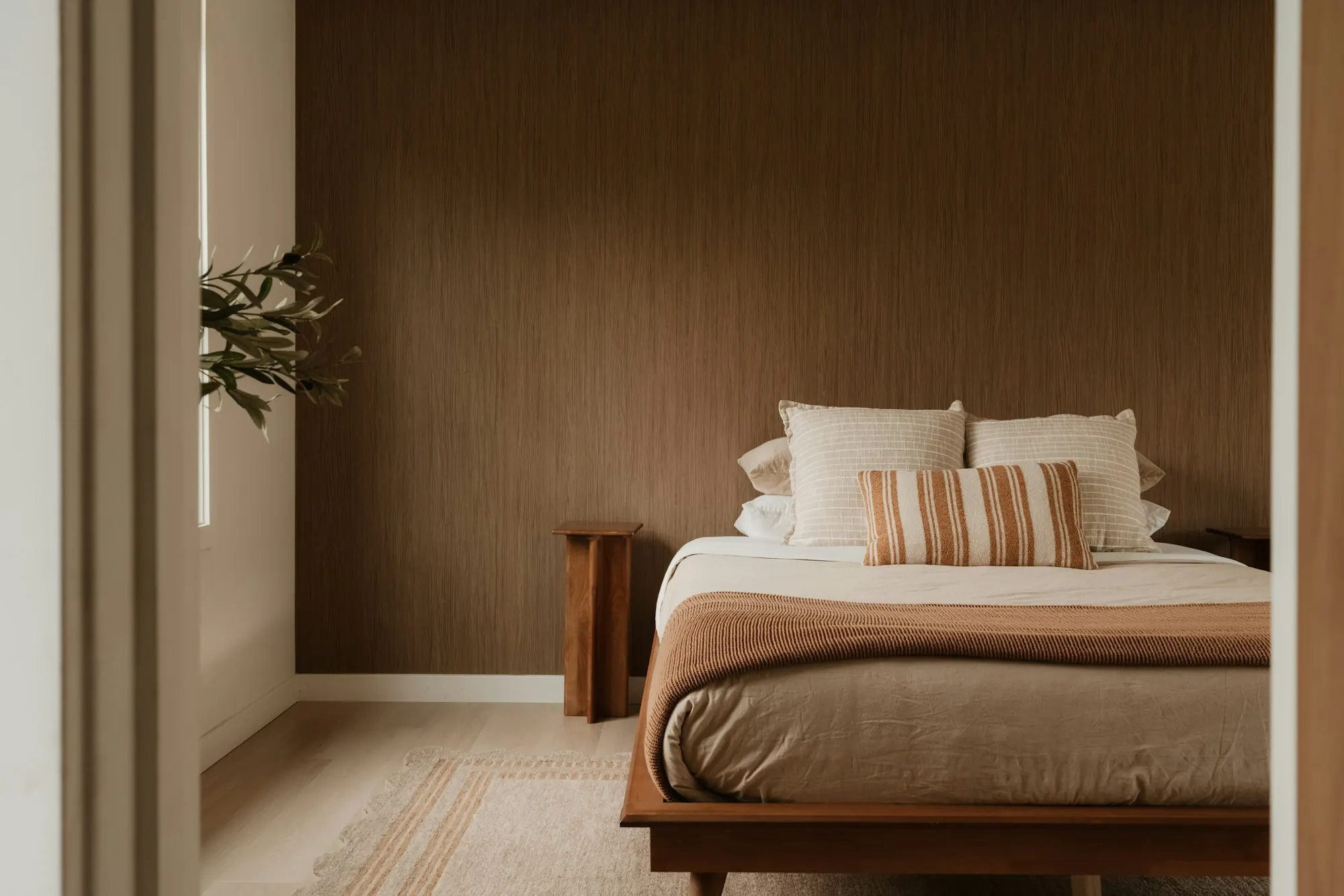 Papier peint brun texturé sur le mur d’une chambre : lit en bois, linge écru, coussin rayé, tapis clair.