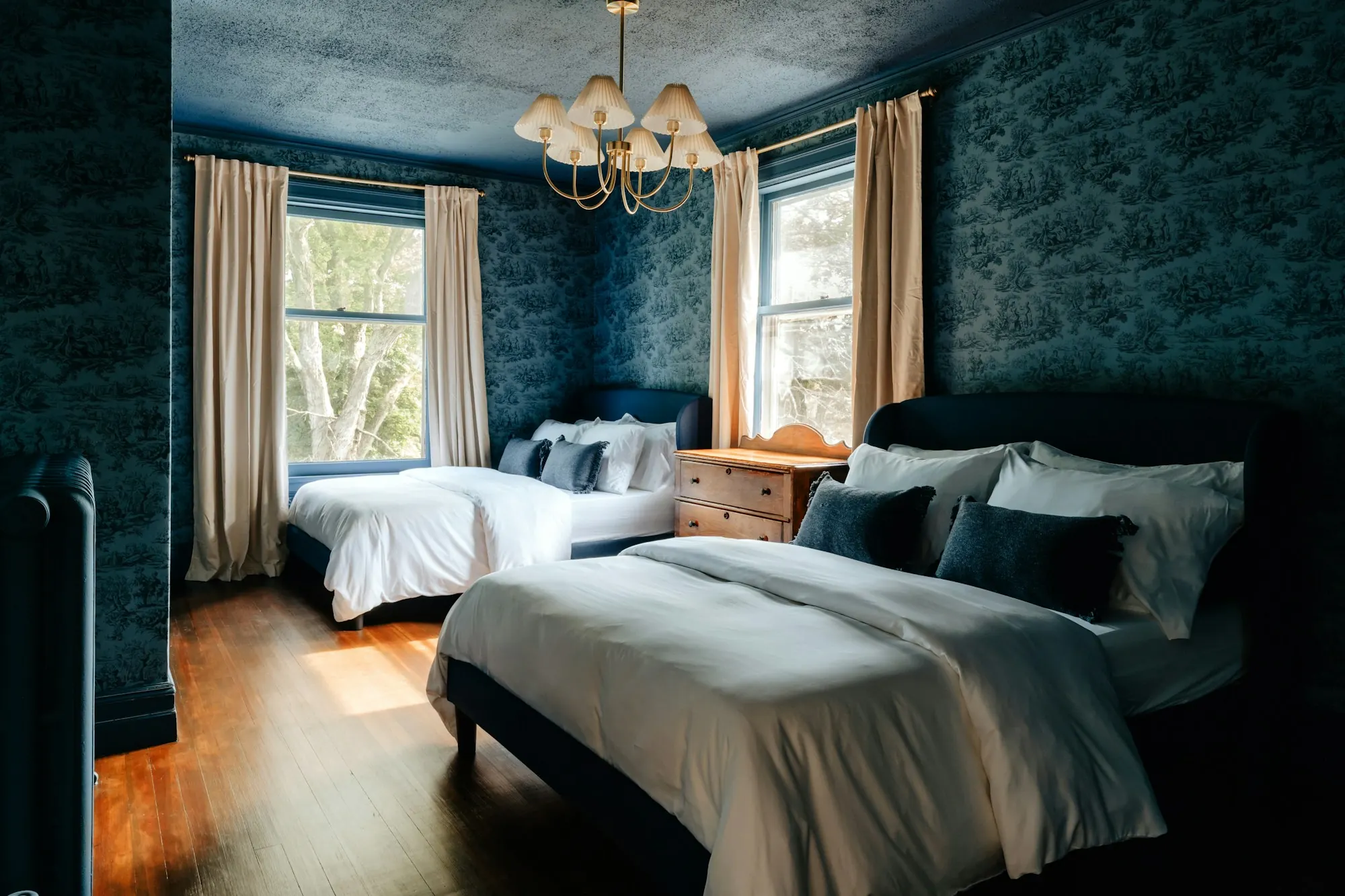 Chambre aux murs et plafond tapissés d’une toile bleu nuit à motifs, deux lits, rideaux beiges, parquet foncé.