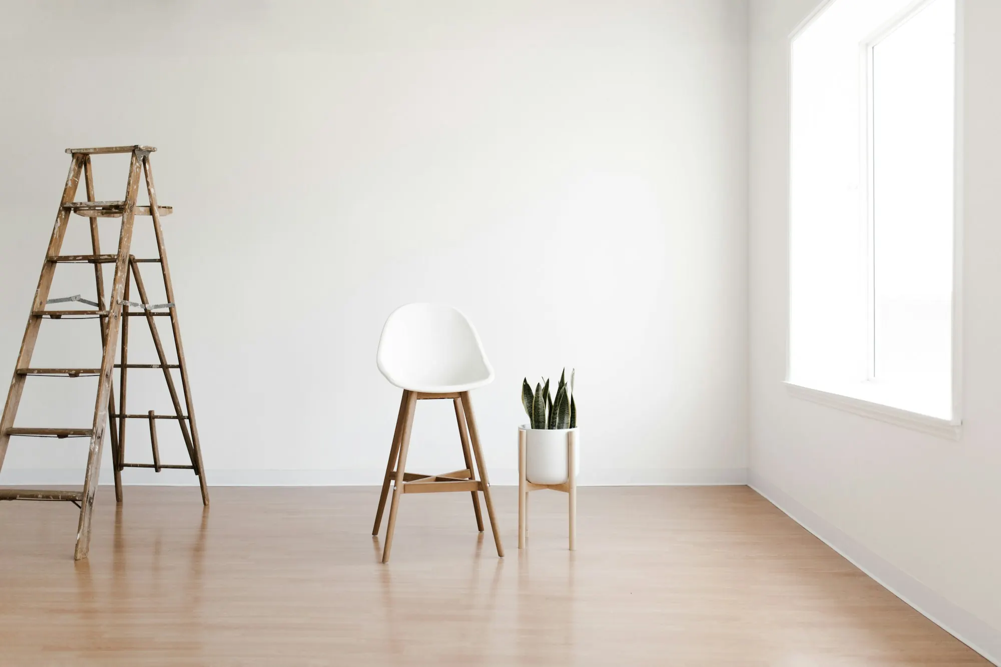 Pièce vide aux murs blancs et sol en bois clair : escabeau en bois, tabouret blanc et plante près d’une fenêtre.