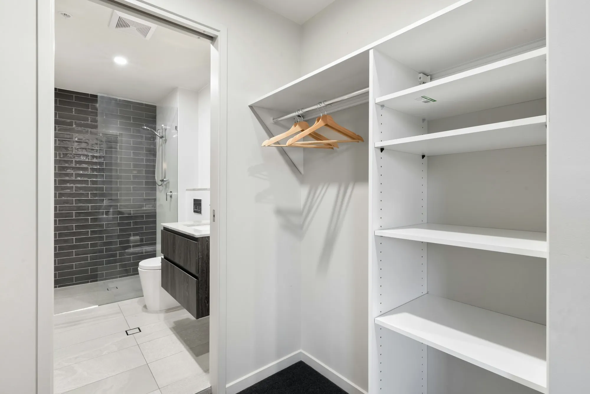 Placard ouvert blanc : étagères réglables et penderie à cintres en bois, salle d’eau visible par la porte.
