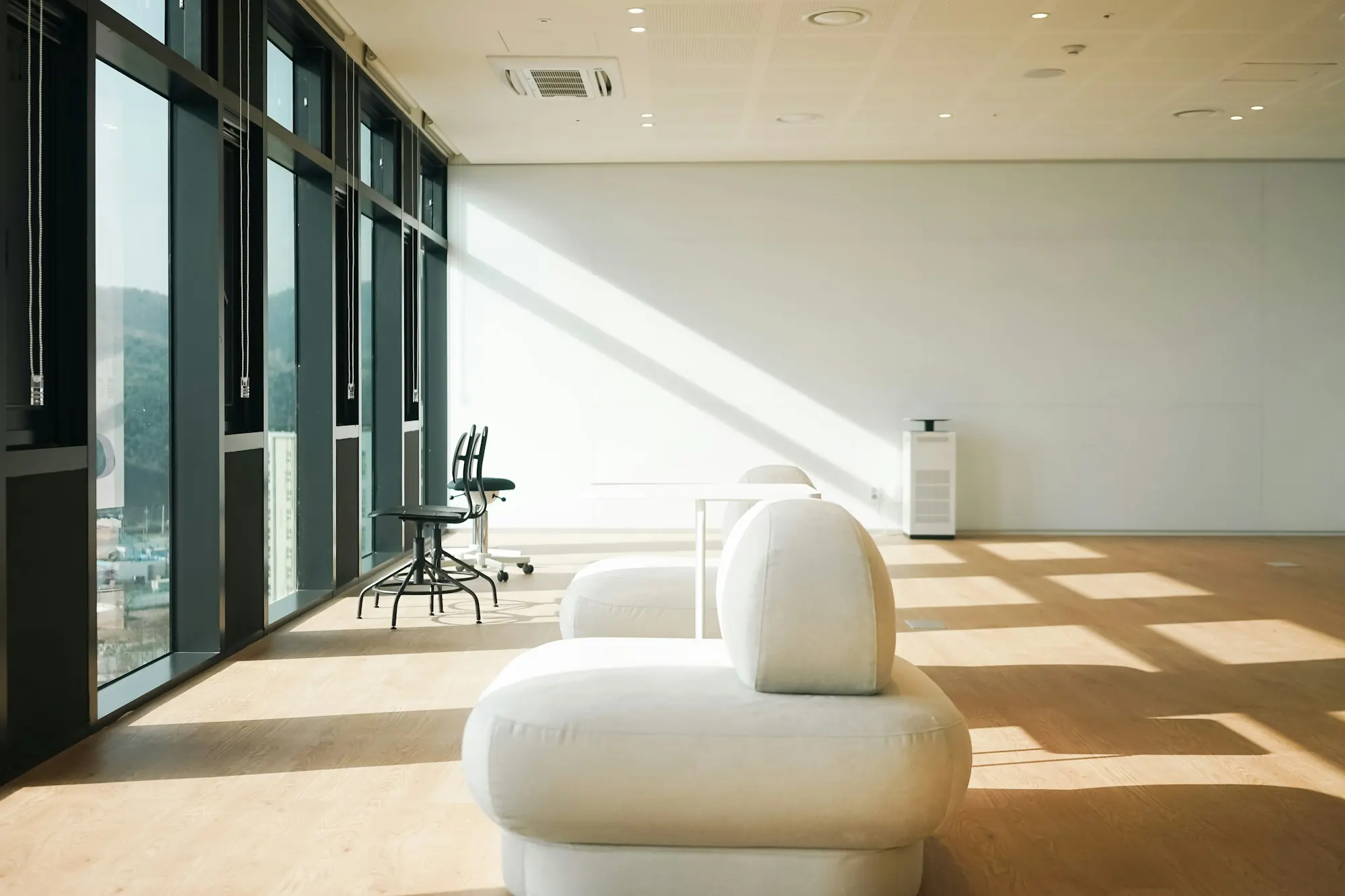 Plateau de bureaux baigné de soleil : sol en bois clair, banquette blanche, tabourets noirs le long de la façade vitrée.