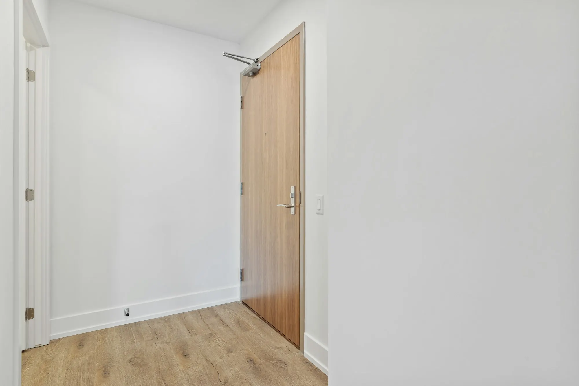 Entrée aux murs blancs : porte en bois clair à ferme-porte, sol d’aspect bois.