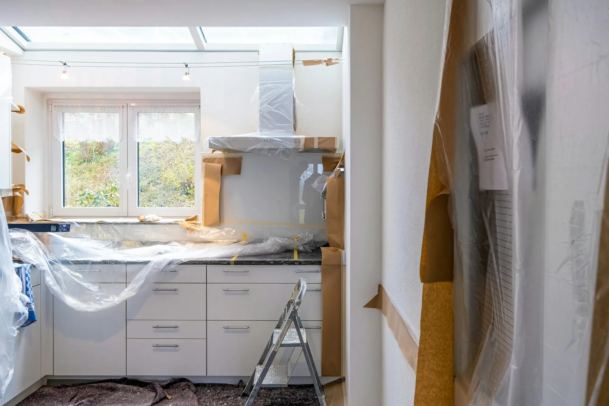 Rénovation de cuisine : façades blanches protégées de films plastique et de papier kraft, escabeau au premier plan.
