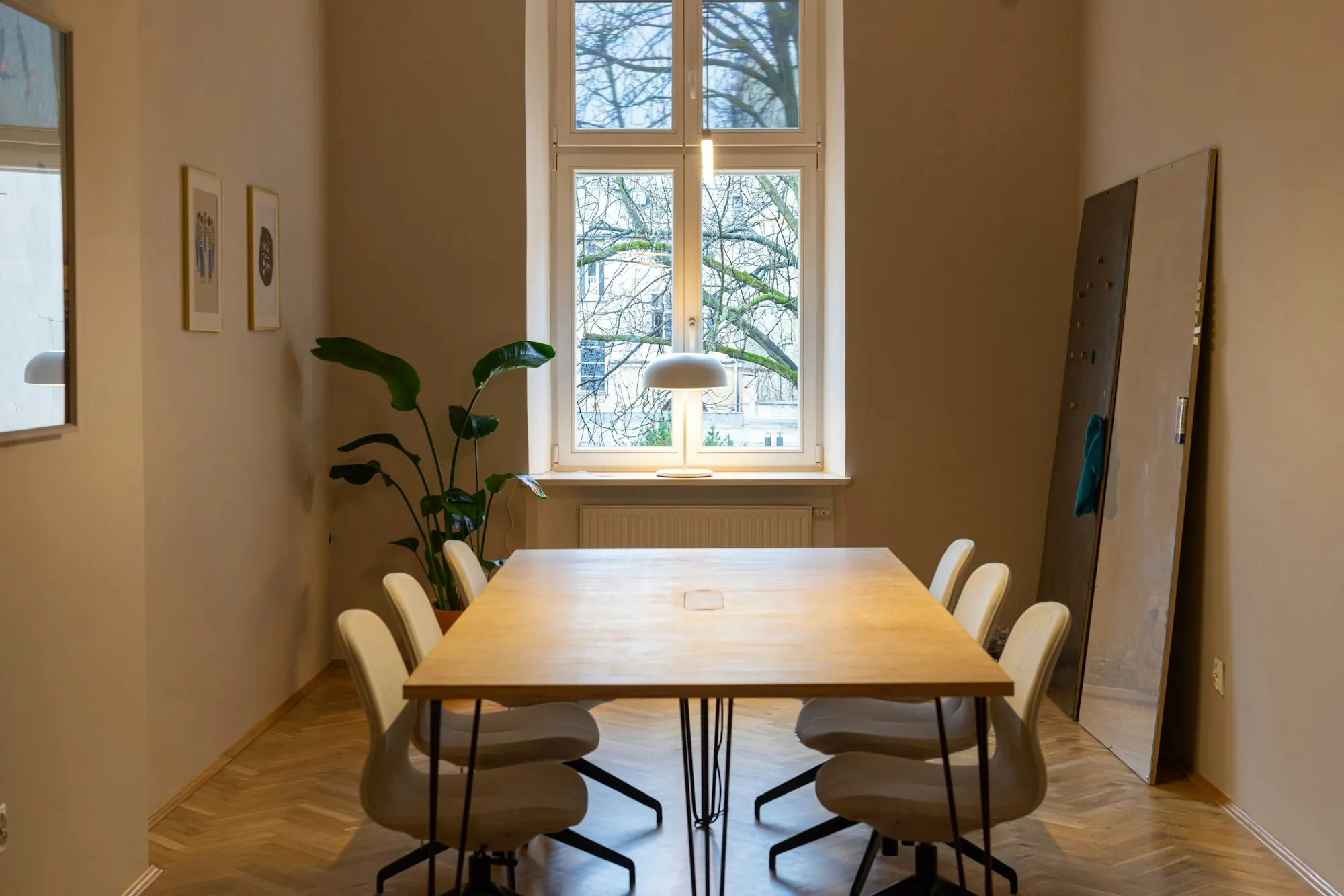 Salle de réunion sur parquet à bâtons rompus : table en bois, chaises blanches, haute fenêtre et grande plante.