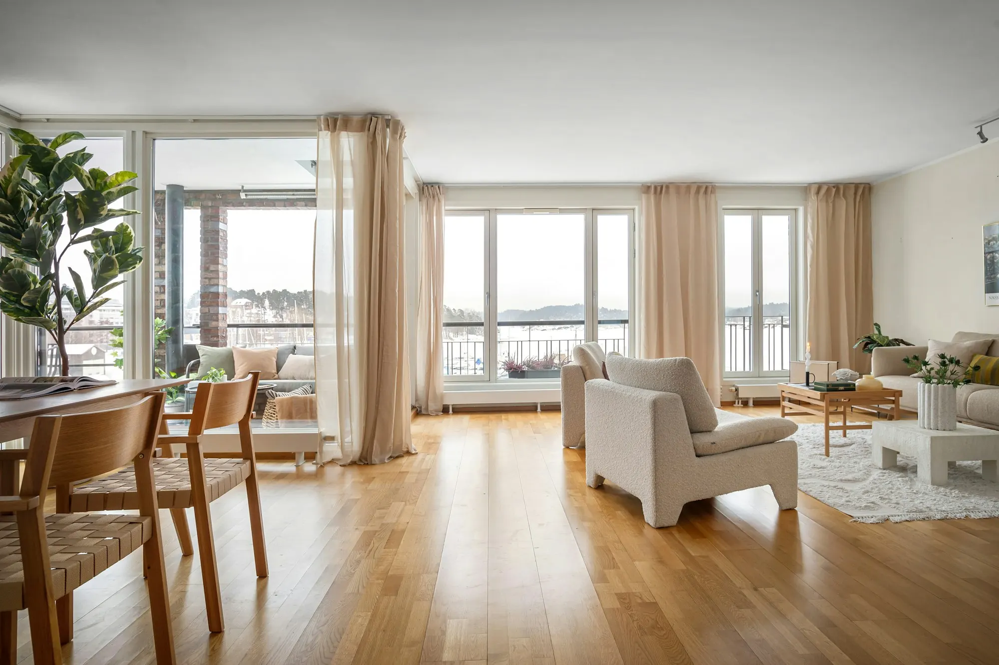 Grand séjour lumineux : parquet en bois blond, voilages écrus devant de larges baies, fauteuils bouclette.