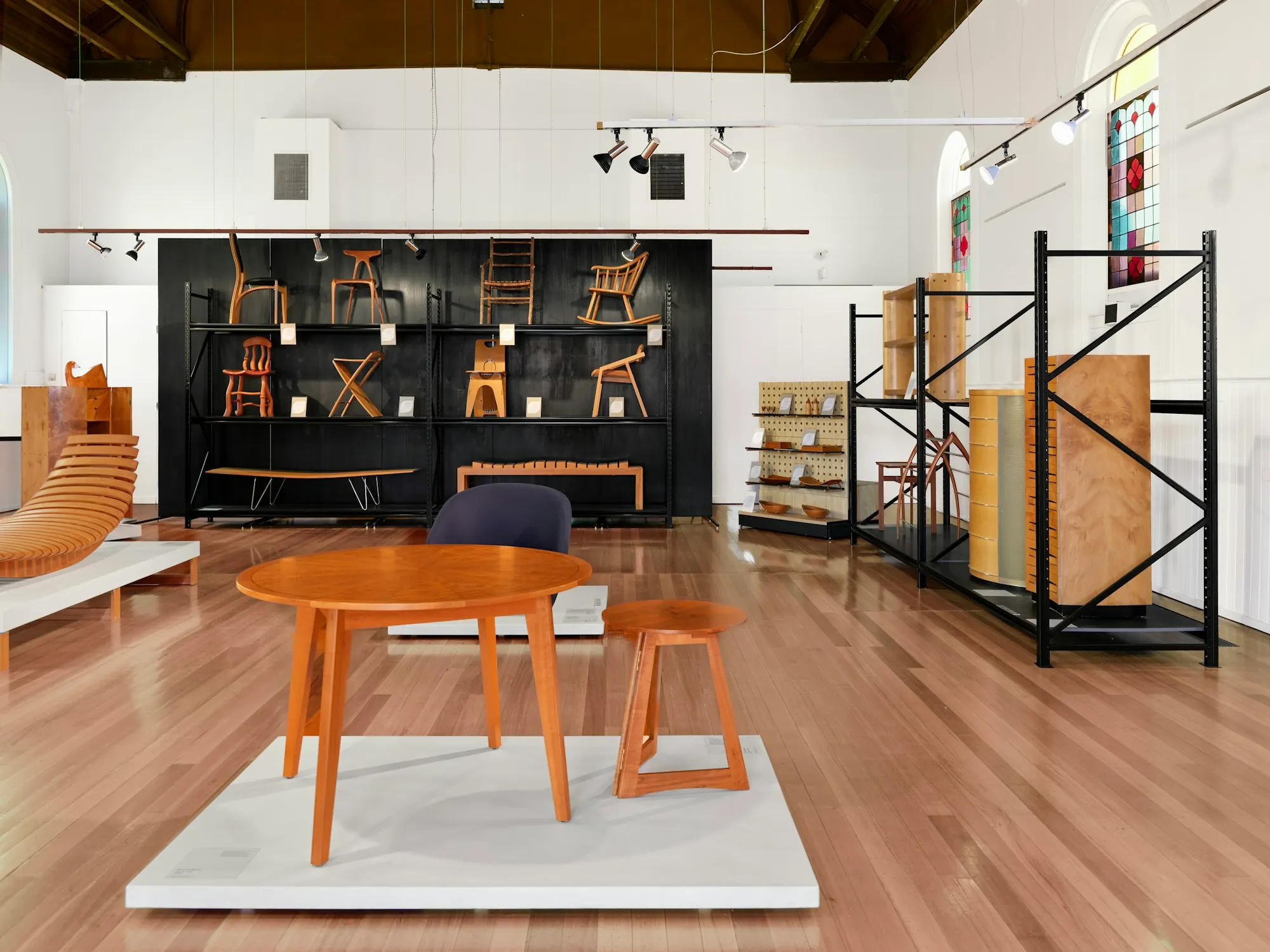 Showroom de mobilier en bois : table et tabourets sur un podium blanc, chaises exposées sur un mur noir, parquet.
