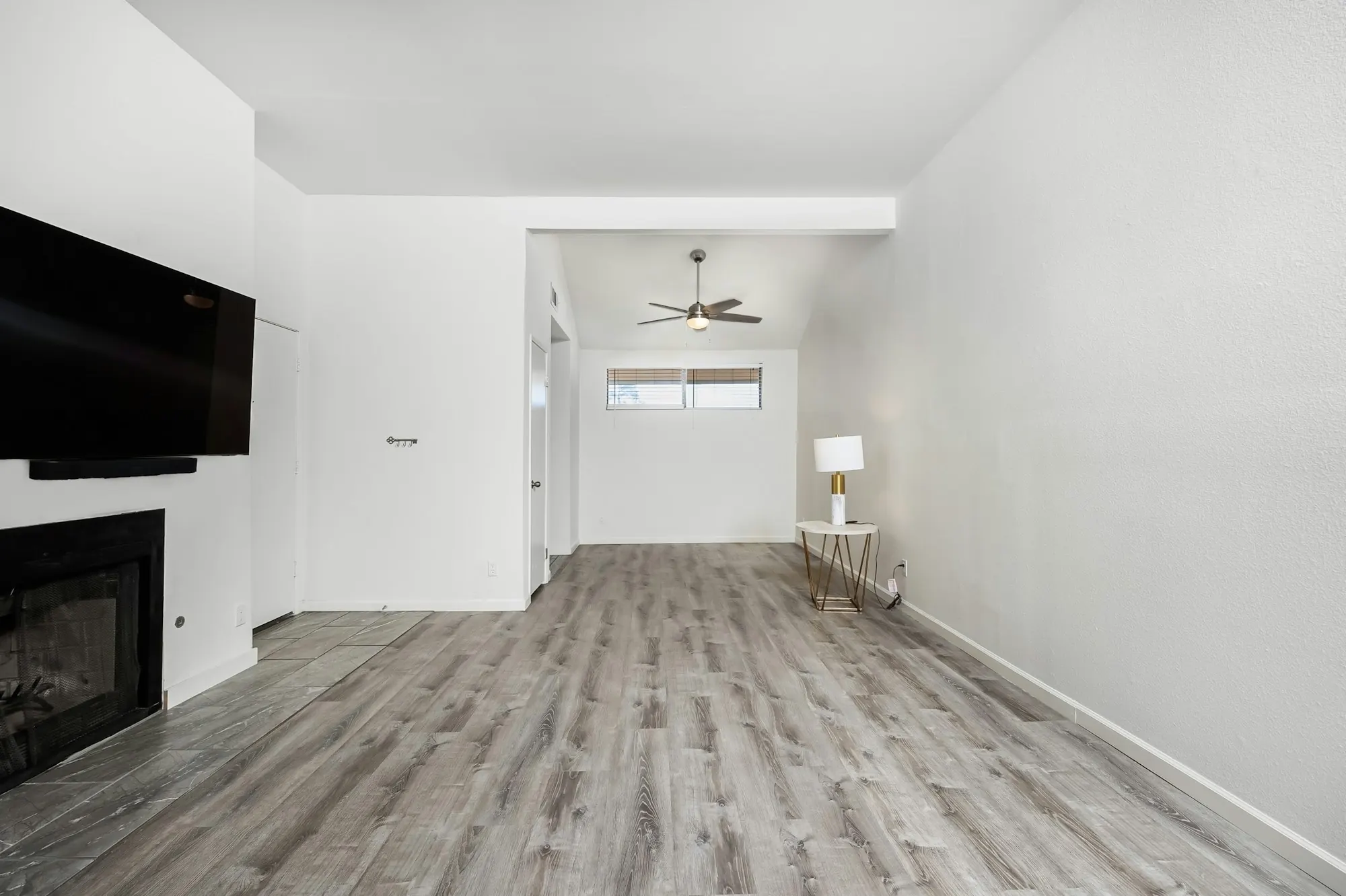 Pièce vide aux murs blancs, sol à lames d’aspect bois gris, cheminée encastrée et ventilateur au plafond.