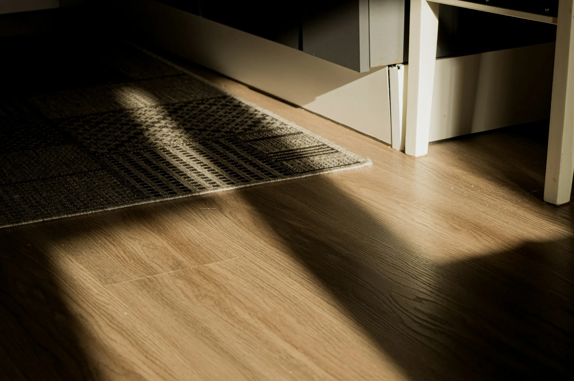 Rayon de soleil sur un sol à lames d’aspect bois clair, bord d’un tapis tissé et d’un meuble blanc.
