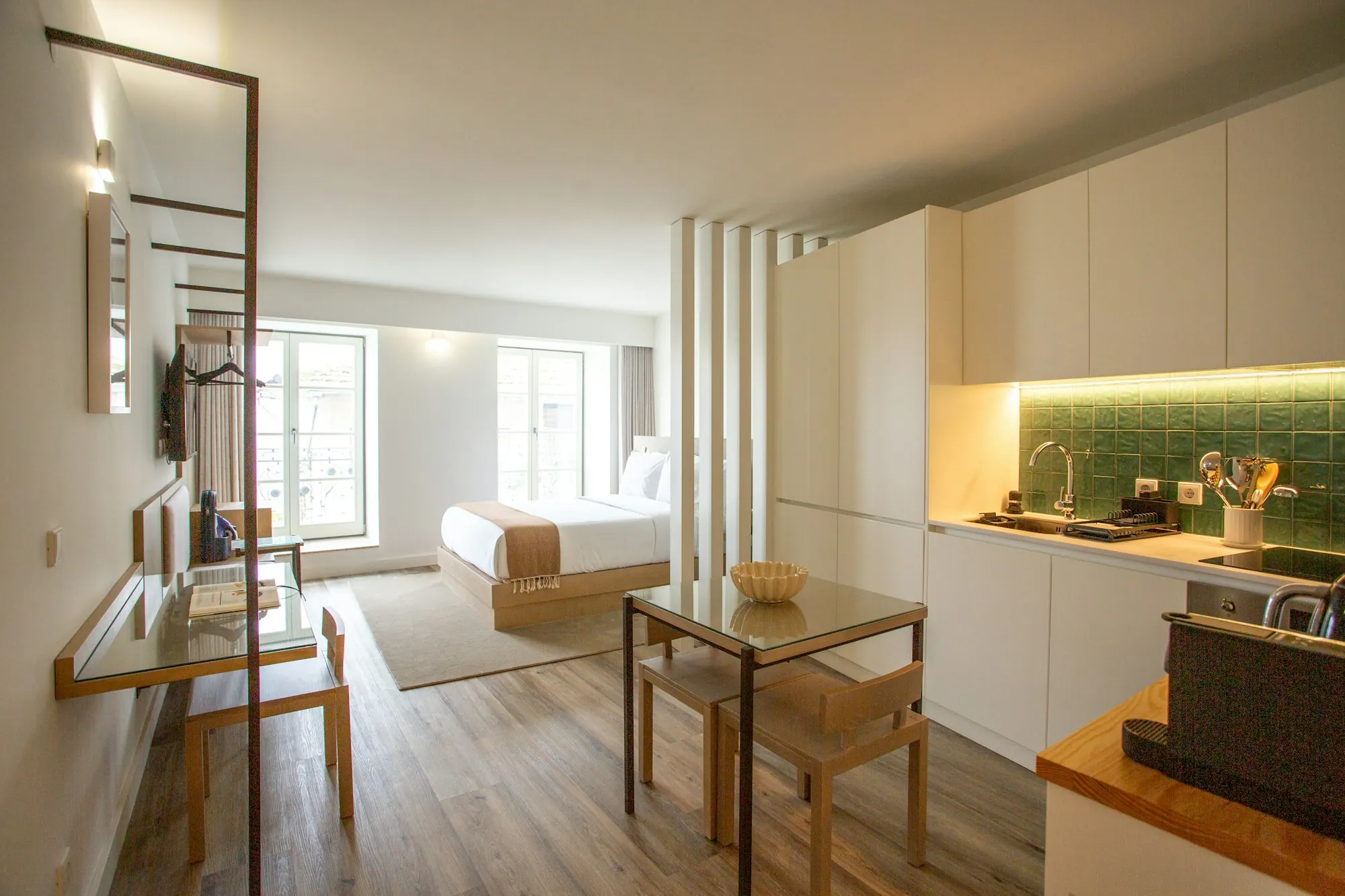 Studio aménagé : lit au fond, kitchenette blanche à crédence verte, petite table et sol d’aspect bois.